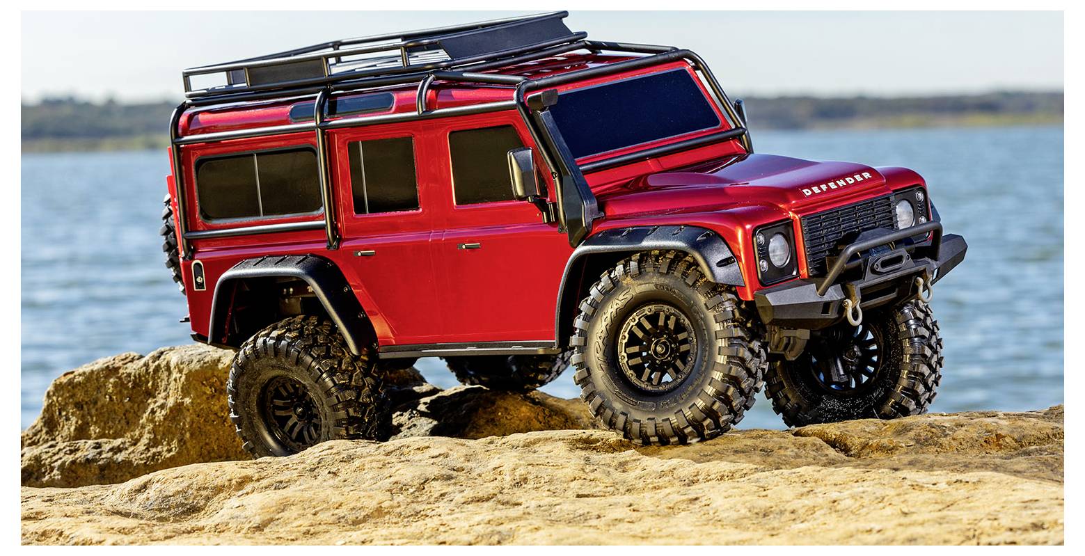 Ein roter ferngesteuerter Geländewagen mit robustem Design sitzt auf Felsen neben einem Gewässer und vermittelt ein Gefühl von Abenteuer.