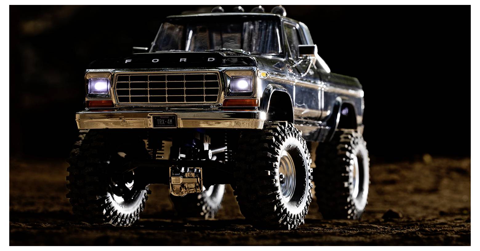 Ein Spielzeug-Lastwagen mit großen Geländereifen und eingeschalteten Scheinwerfern, der nachts auf einer Schmutzoberfläche geparkt ist.