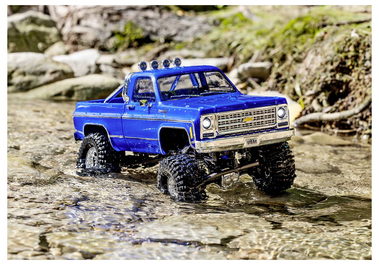 Ein blauer Spielzeug-Geländewagen mit Offroad-Reifen fährt durch einen seichten Bach, umgeben von Steinen und Vegetation.