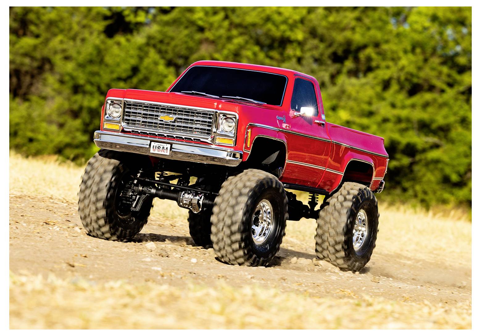Ein roter Monstertruck mit großen überdimensionalen Reifen fährt auf einem Erdweg in einem offenen Feld, umgeben von grünen Bäumen im Hintergrund.