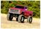 Ein roter Monstertruck mit großen überdimensionalen Reifen fährt auf einem Erdweg in einem offenen Feld, umgeben von grünen Bäumen im Hintergrund.