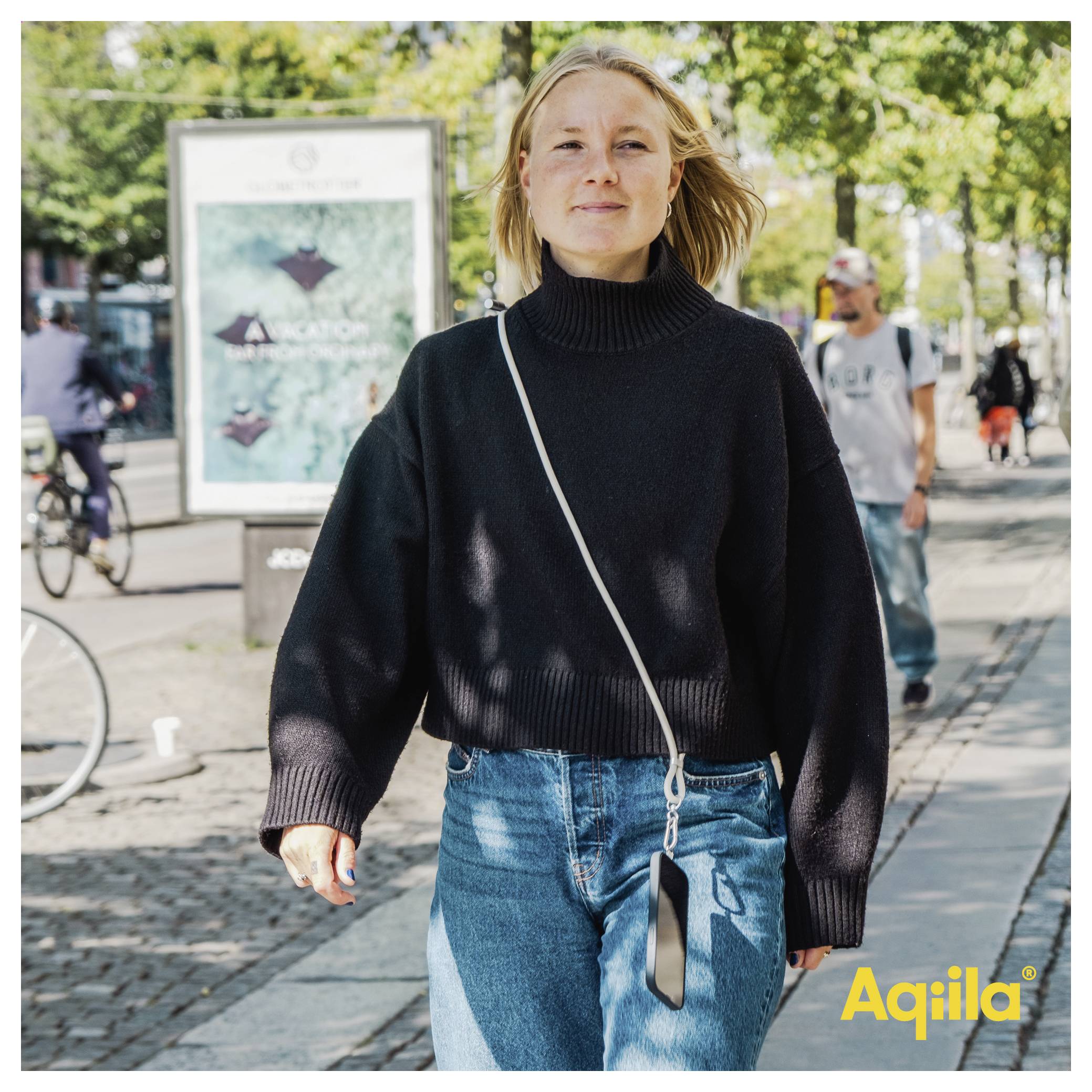 Eine Person geht an einem sonnigen Tag auf einer Stadtstraße, trägt einen schwarzen Pullover und Jeans. Im Hintergrund sind Fußgänger und Radfahrer zu sehen.