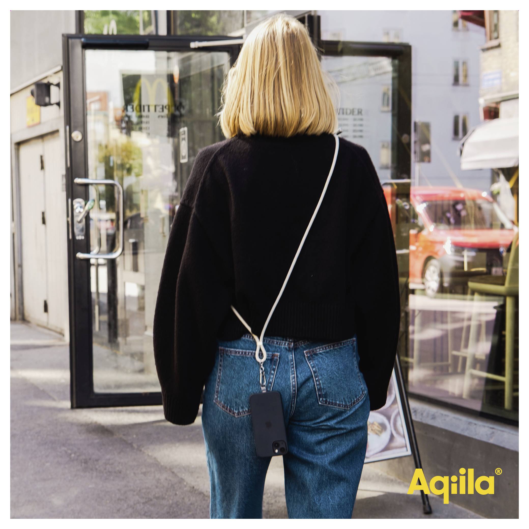 Eine Person mit blonden Haaren nähert sich einer Glastür an einer Stadtstraße, trägt einen schwarzen Pullover und Jeans, mit einem Telefon an einem Umhängeriemen.