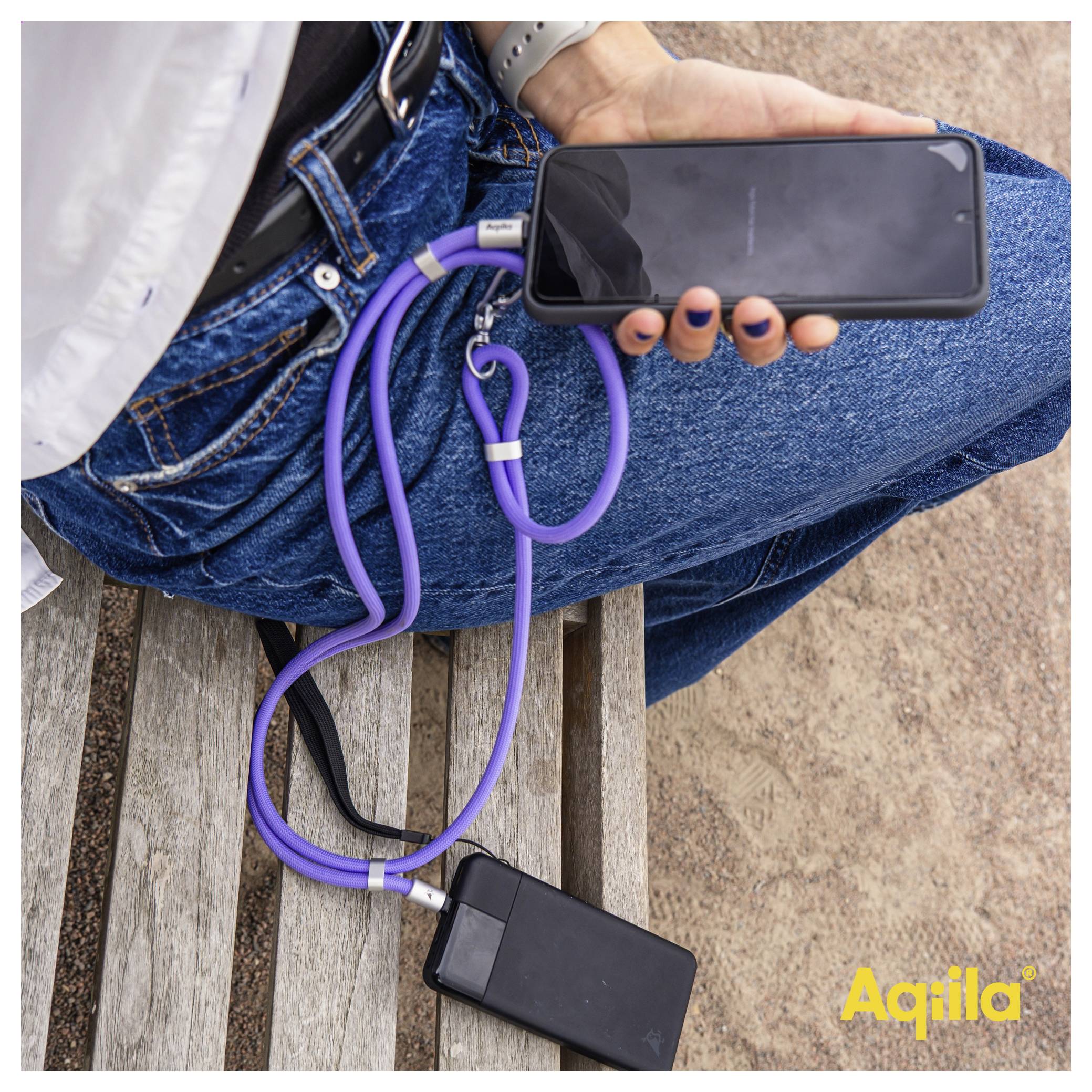 Eine Person sitzt auf einer Bank und hält ein Smartphone, das an einem violetten Trageriemen befestigt ist und mit einer Powerbank verbunden ist. Die Marke 'Aqila' ist sichtbar.