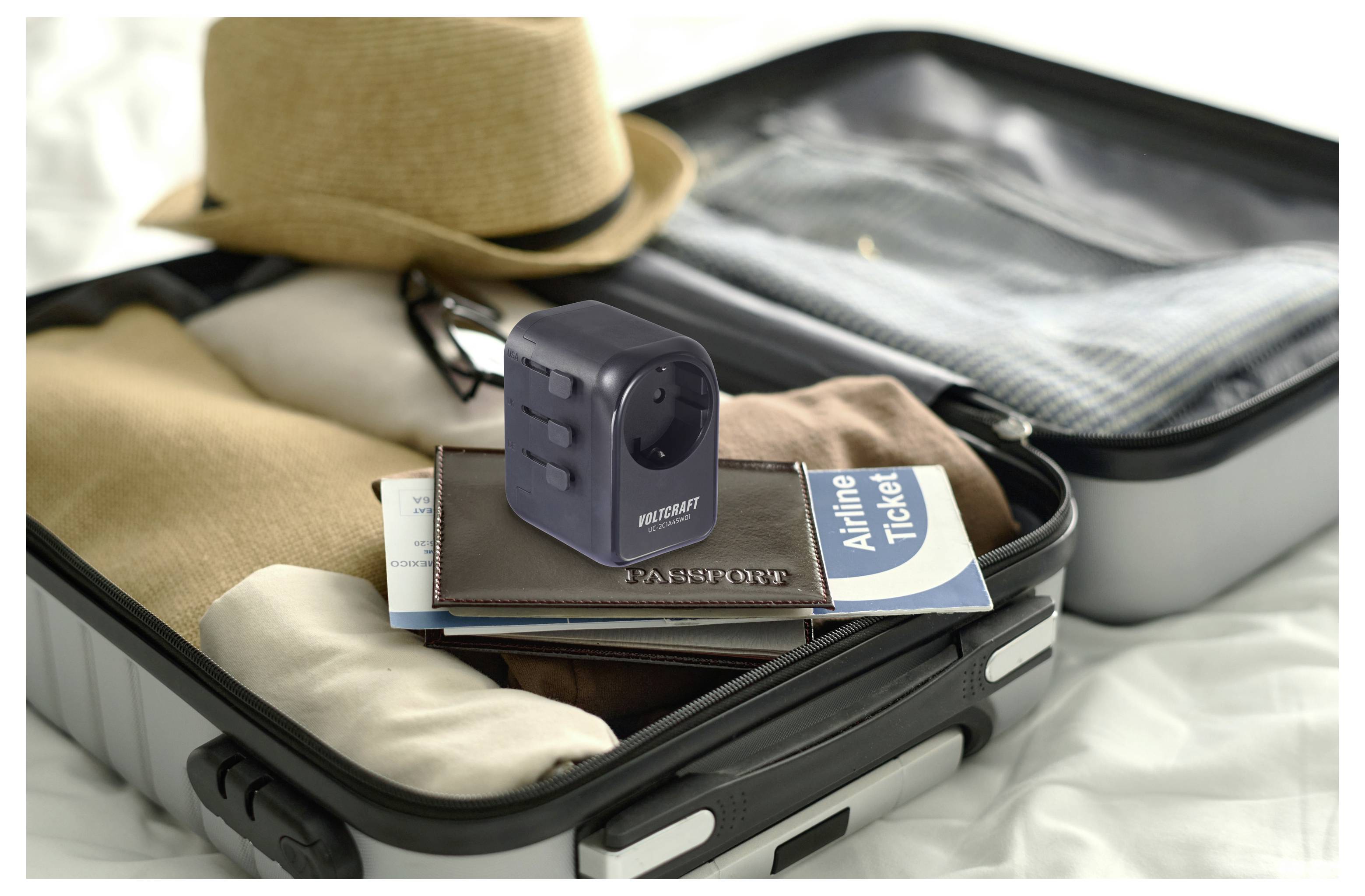 Ein offener Koffer auf einem Bett enthält Kleidung, einen Reisepass, ein Flugticket und einen universellen Reiseadapter, mit einem Hut, der oben aufliegt.