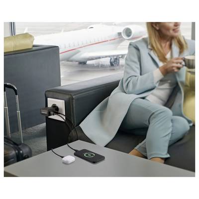 Eine Person in einem türkisfarbenen Outfit sitzt in einer Flughafenlounge und trinkt Kaffee. Ein Telefon lädt drahtlos auf einem Tisch auf, mit einem Flugzeug draußen.