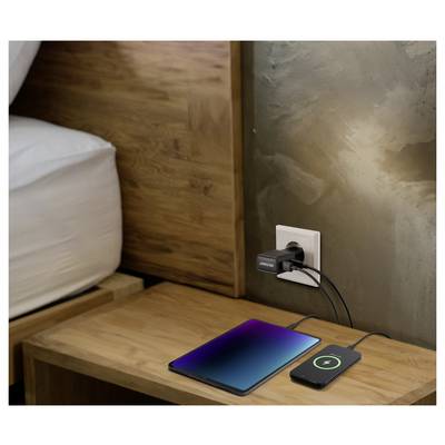 Eine Tablet und ein Smartphone werden auf einem Holznachttisch mit einem Dual-USB-Adapter aufgeladen, der in eine Steckdose neben einem Bett eingesteckt ist.