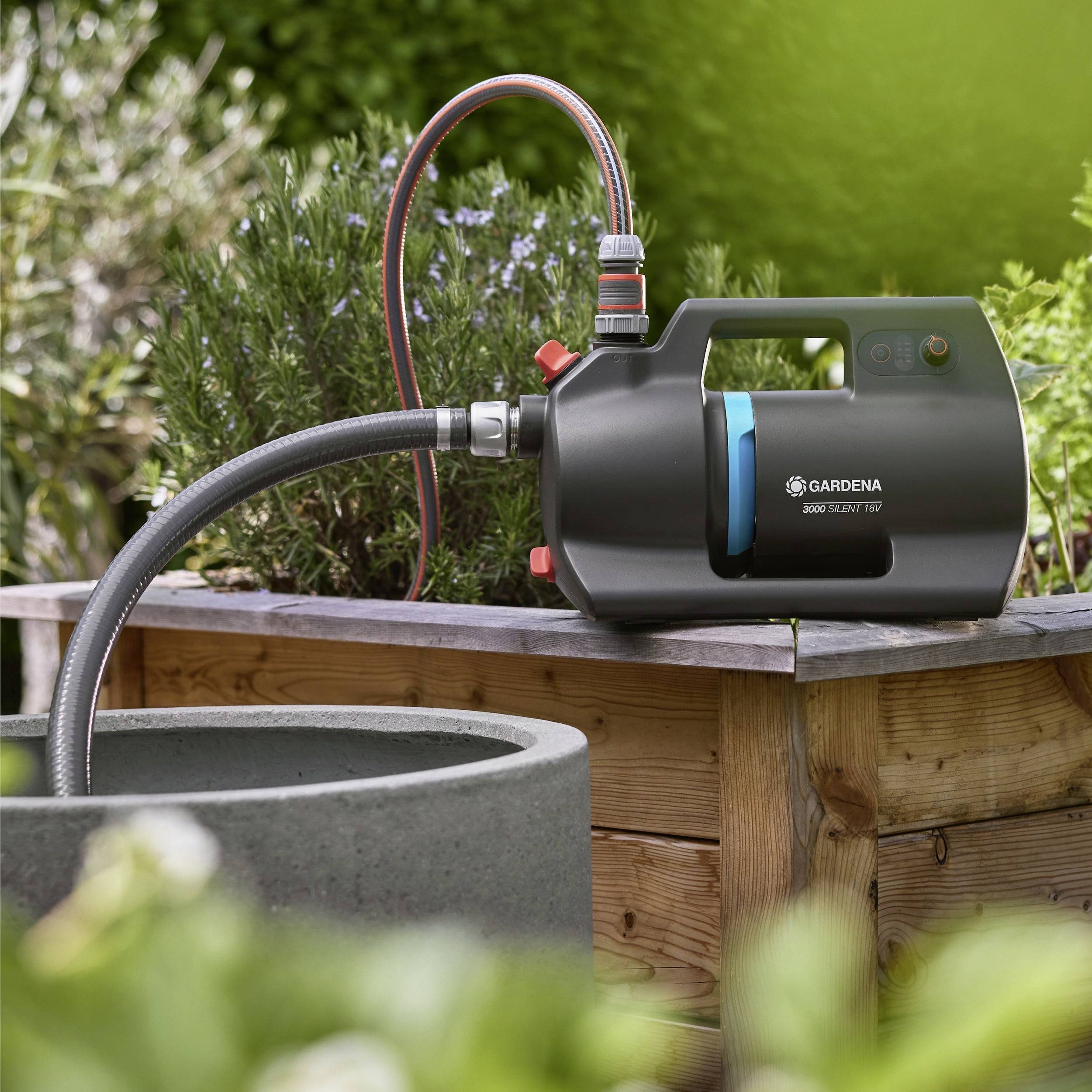 Eine schwarze Gardena Wasserpumpe, die an einen Gartenschlauch angeschlossen ist, steht neben einem kreisförmigen steinernen Wasserbecken, umgeben von grünen Pflanzen.