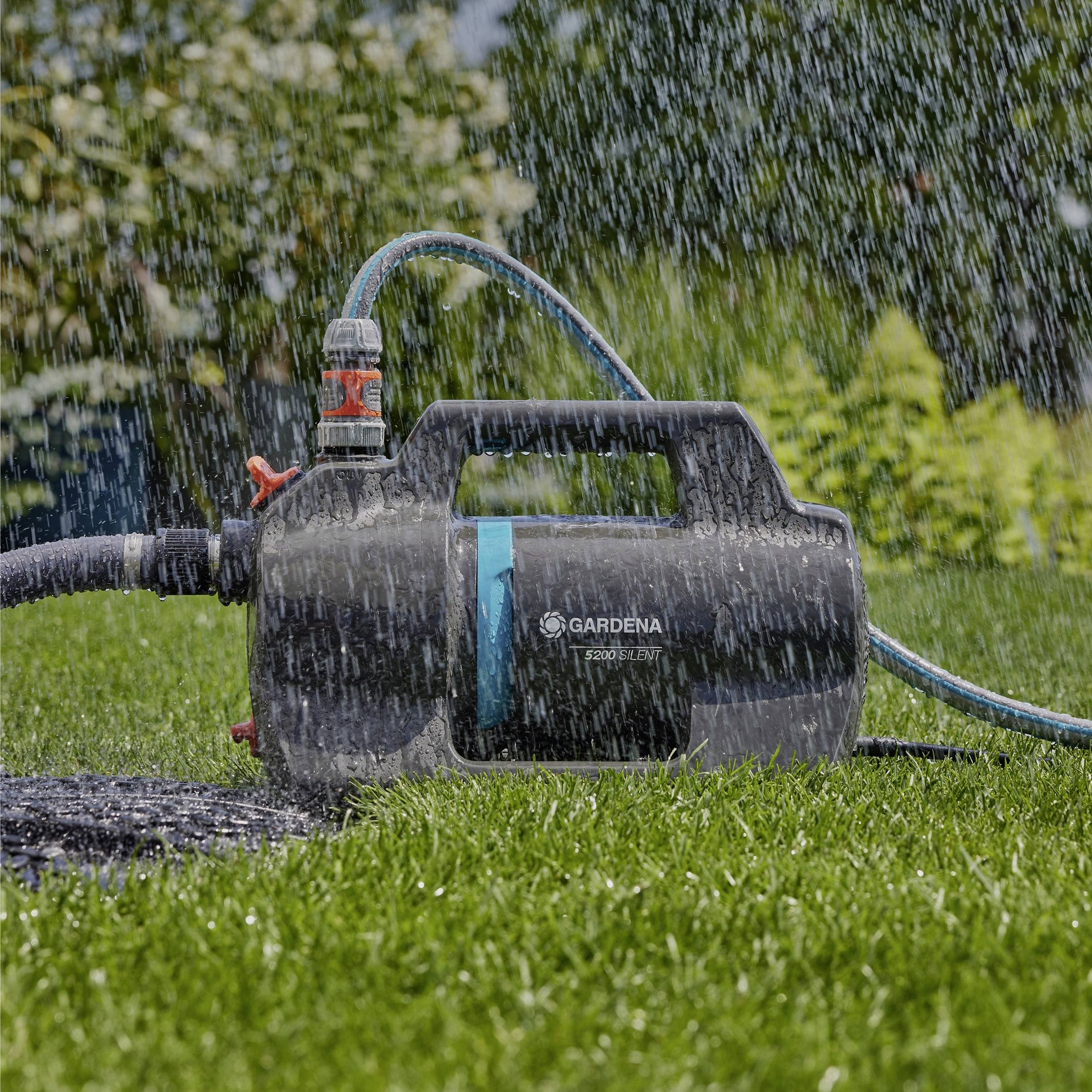 Ein Gartenschlauch-Sprinklersystem bewässert einen grünen Rasen, wobei eine schwarze und blaue Zentrale mit der Aufschrift „Gardena" in Betrieb ist und von Grünpflanzen umgeben ist.