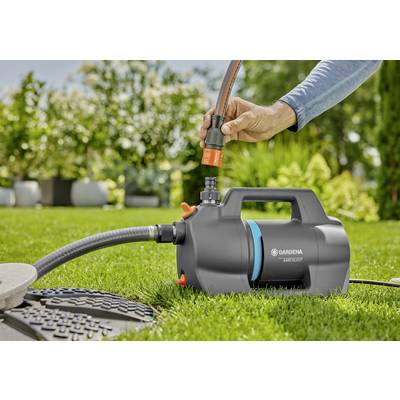 Eine Person verbindet einen Schlauch mit einer Gardena-Gartenpumpe auf einem Rasen, umgeben von Grünflächen und Blumenkübeln in einer sonnigen Außenumgebung.
