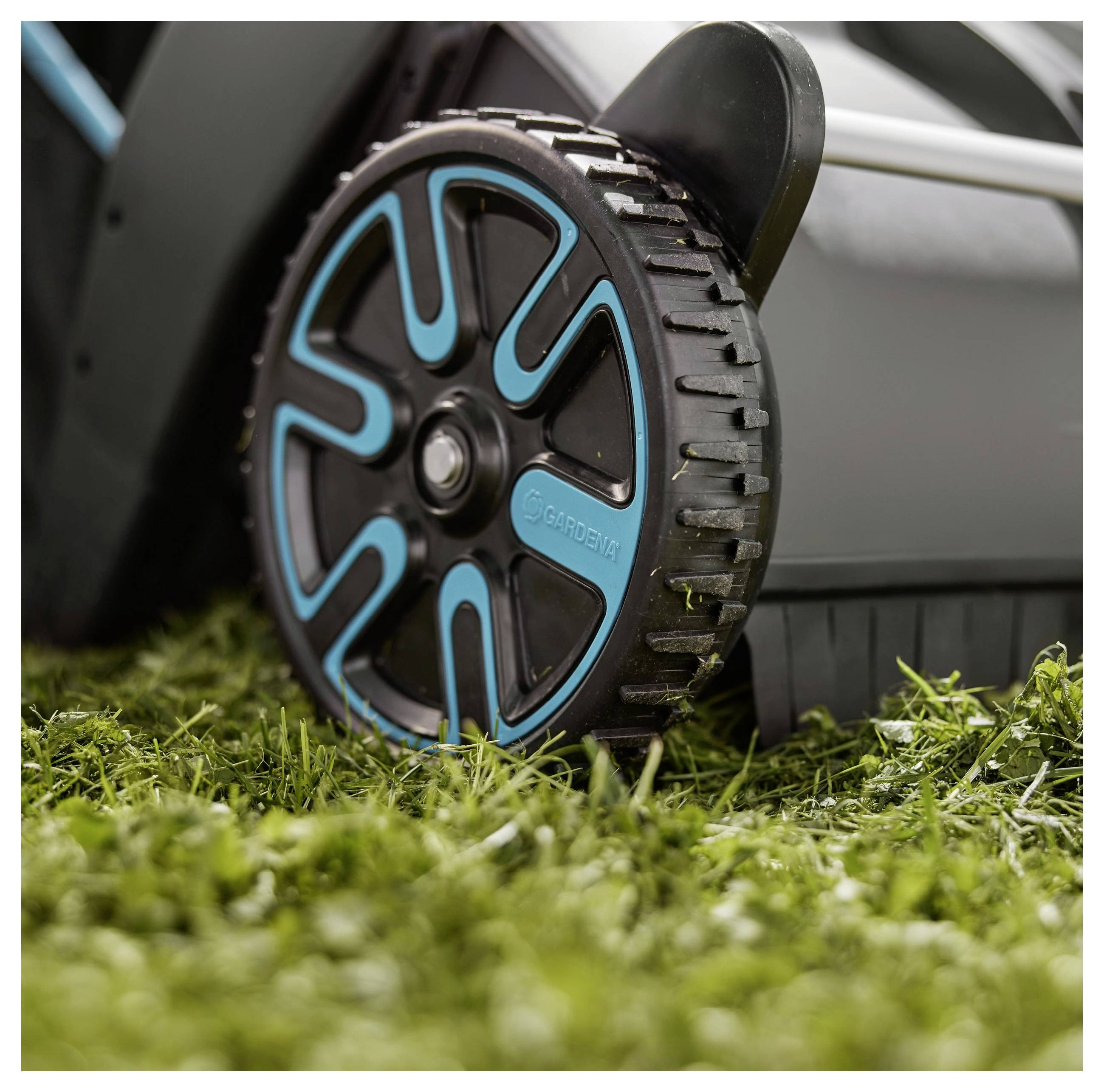 Nahaufnahme eines Rasenmäherades auf Gras. Das Rad hat ein schwarzblaues Design mit sichtbarem Profil und zeigt die Marke „Gardena".