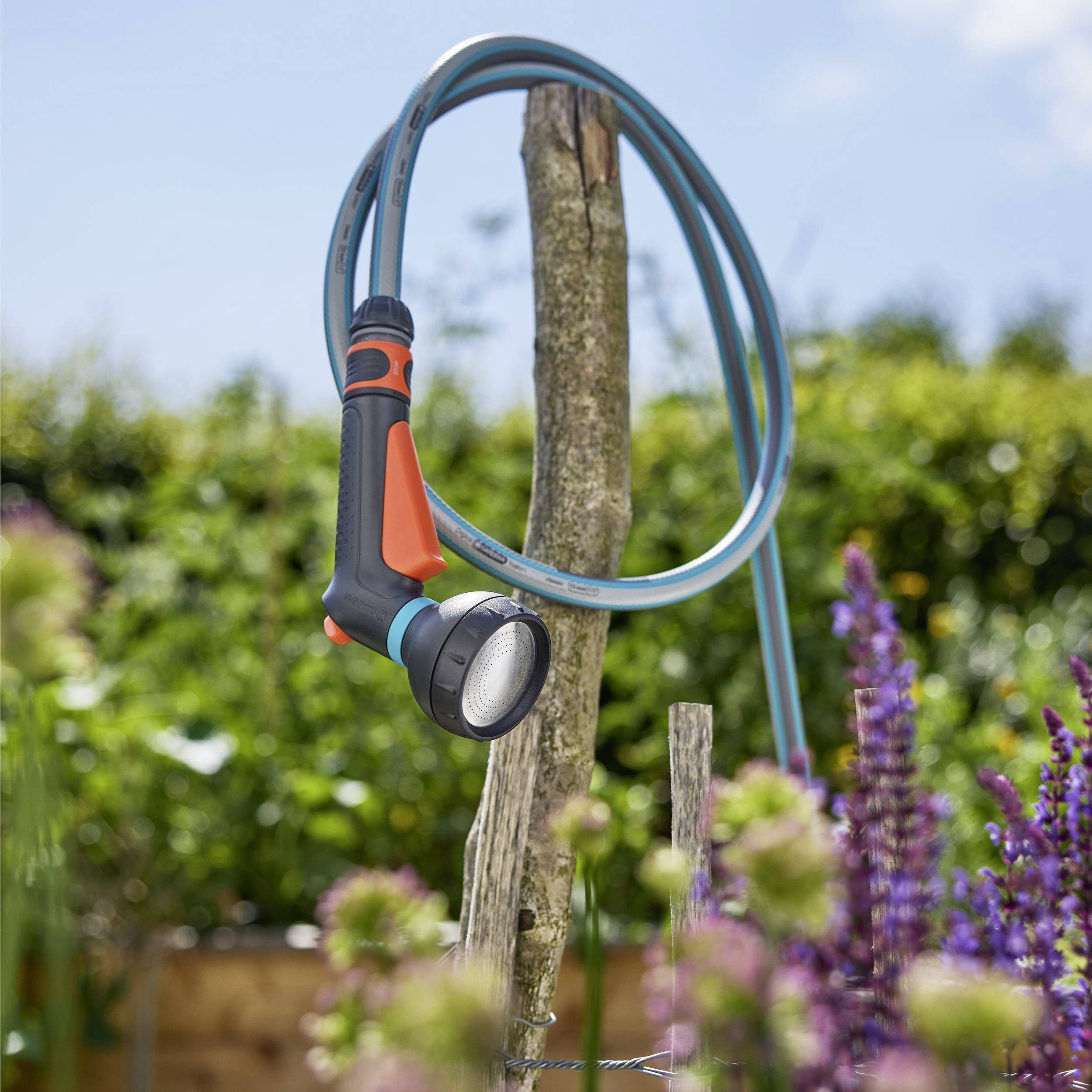 Eine Gartenschlauchbrause, die an einem Holzpfosten befestigt ist, umgeben von verschwommenen Blumen und Grünpflanzen, was eine Hinterhofgartenszene andeutet.