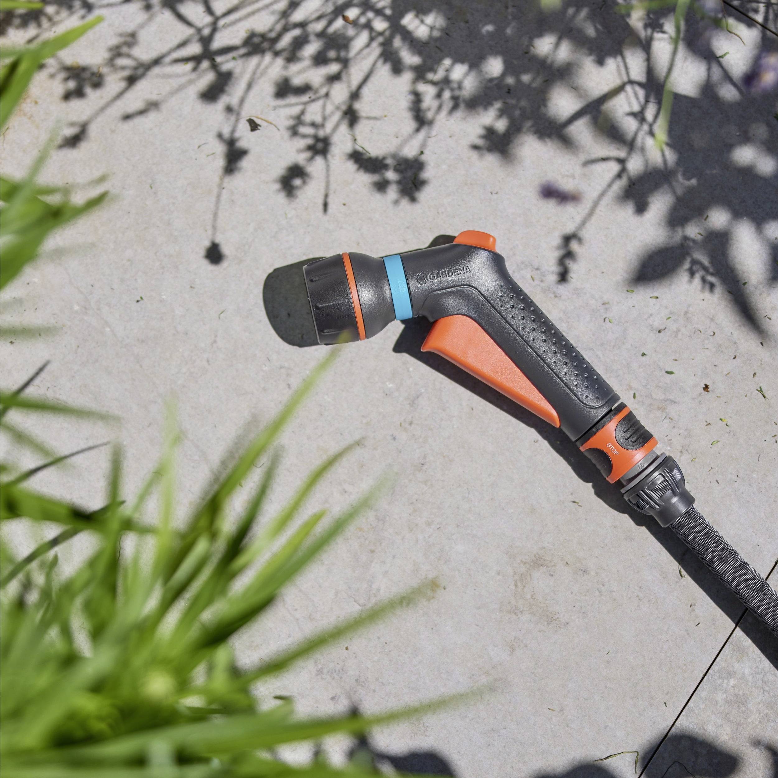 Ein Gartenschlauch mit einer verstellbaren Sprühdüse liegt auf einer gepflasterten Oberfläche, umgeben von Pflanzen, die Schatten auf den Boden werfen.