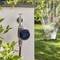 Eine Gartenwand-Wasserzeitschaltuhr steuert einen Schlauch in einem sonnigen Garten mit Grünpflanzen. Ein Sprinkler bewässert im Hintergrund den Rasen.