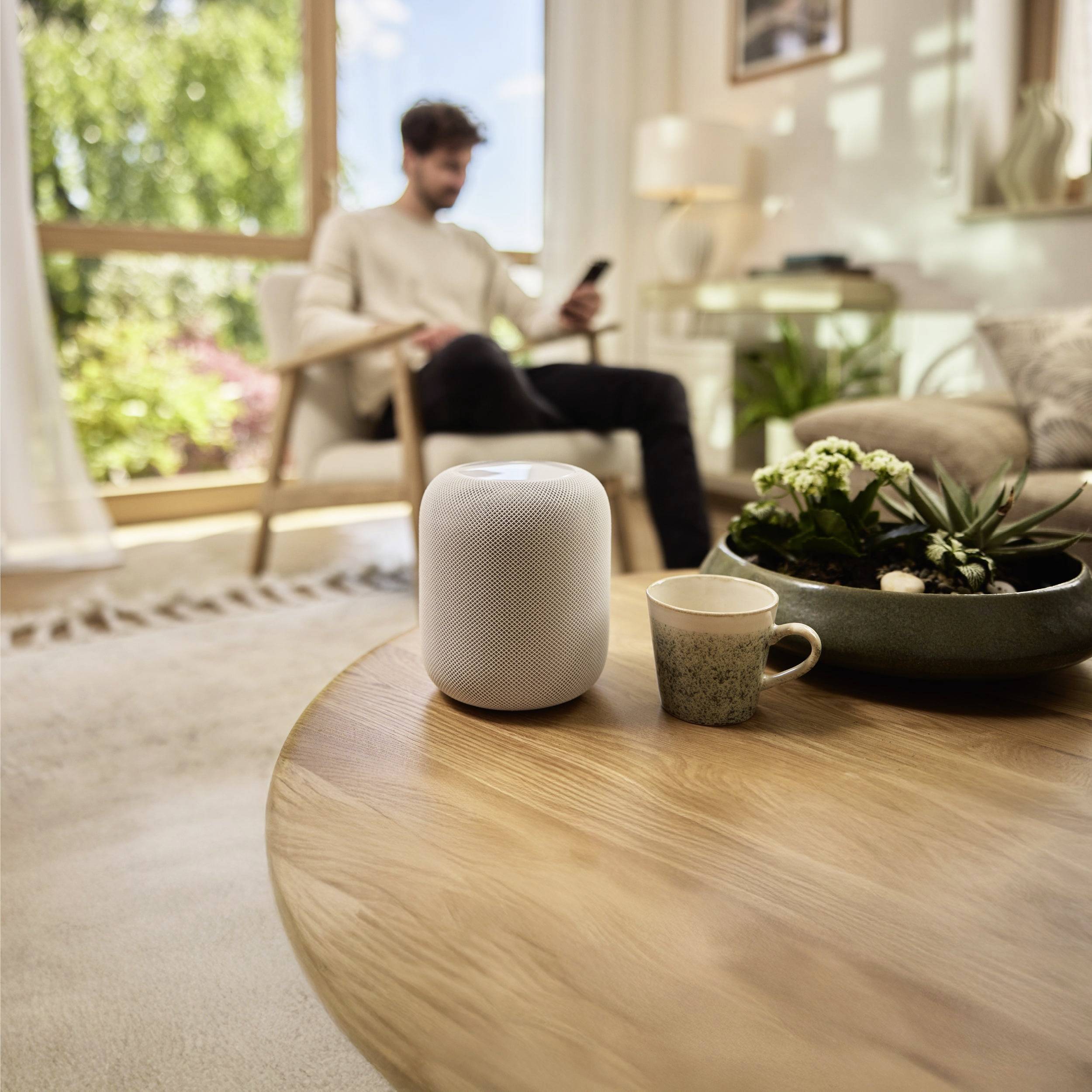 Eine Person sitzt in einem Stuhl und benutzt ein Smartphone in der Nähe eines Fensters, durch das Sonnenlicht hereinströmt. Auf einem Tisch im Vordergrund befinden sich ein Lautsprecher, eine Kaffeetasse und eine Pflanze.