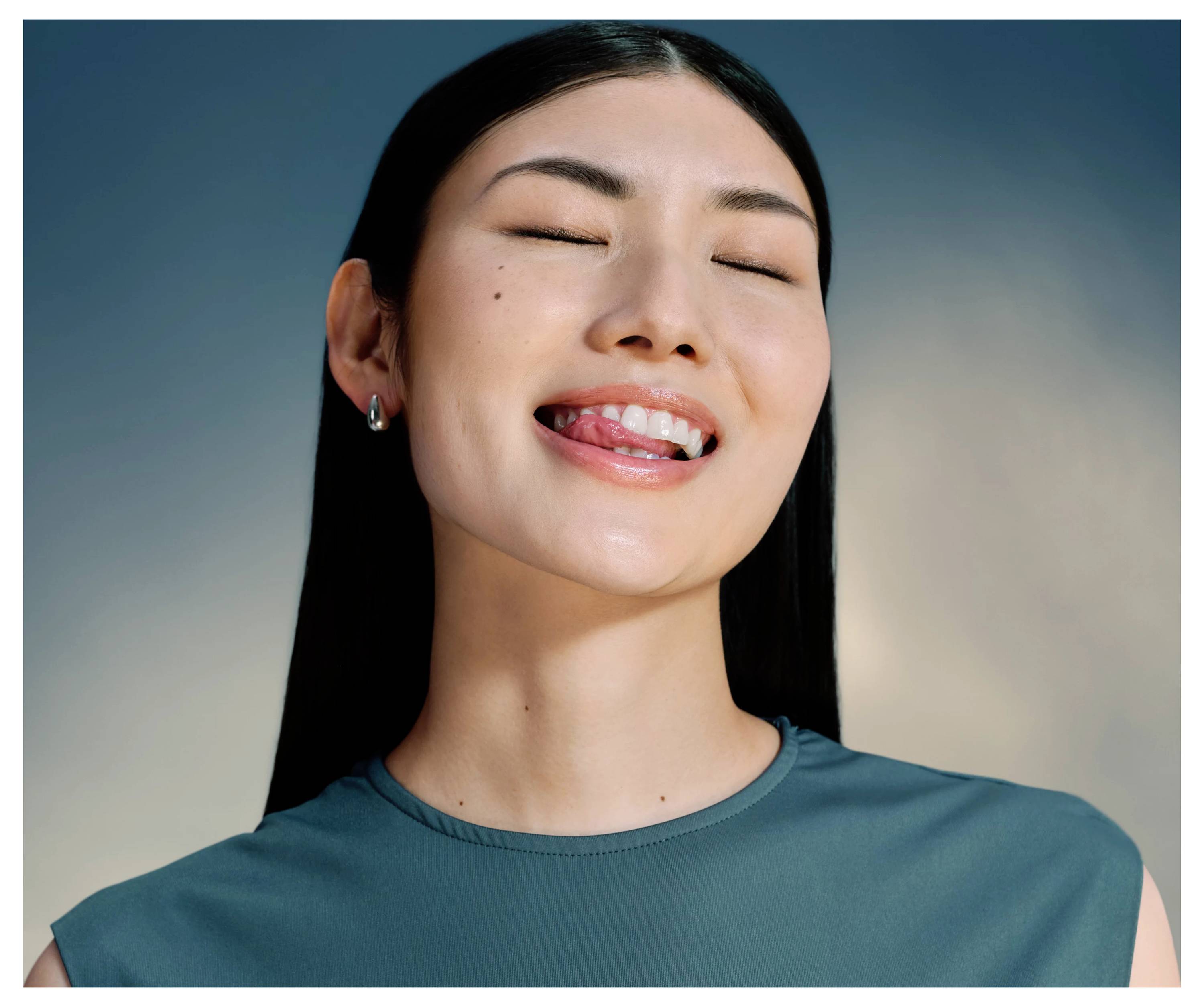 Eine Frau mit langen dunklen Haaren lächelt und streckt verspielt die Zunge heraus, gekleidet in einem petrolfarben ärmellosen Top, vor einem Verlaufshintergrund.