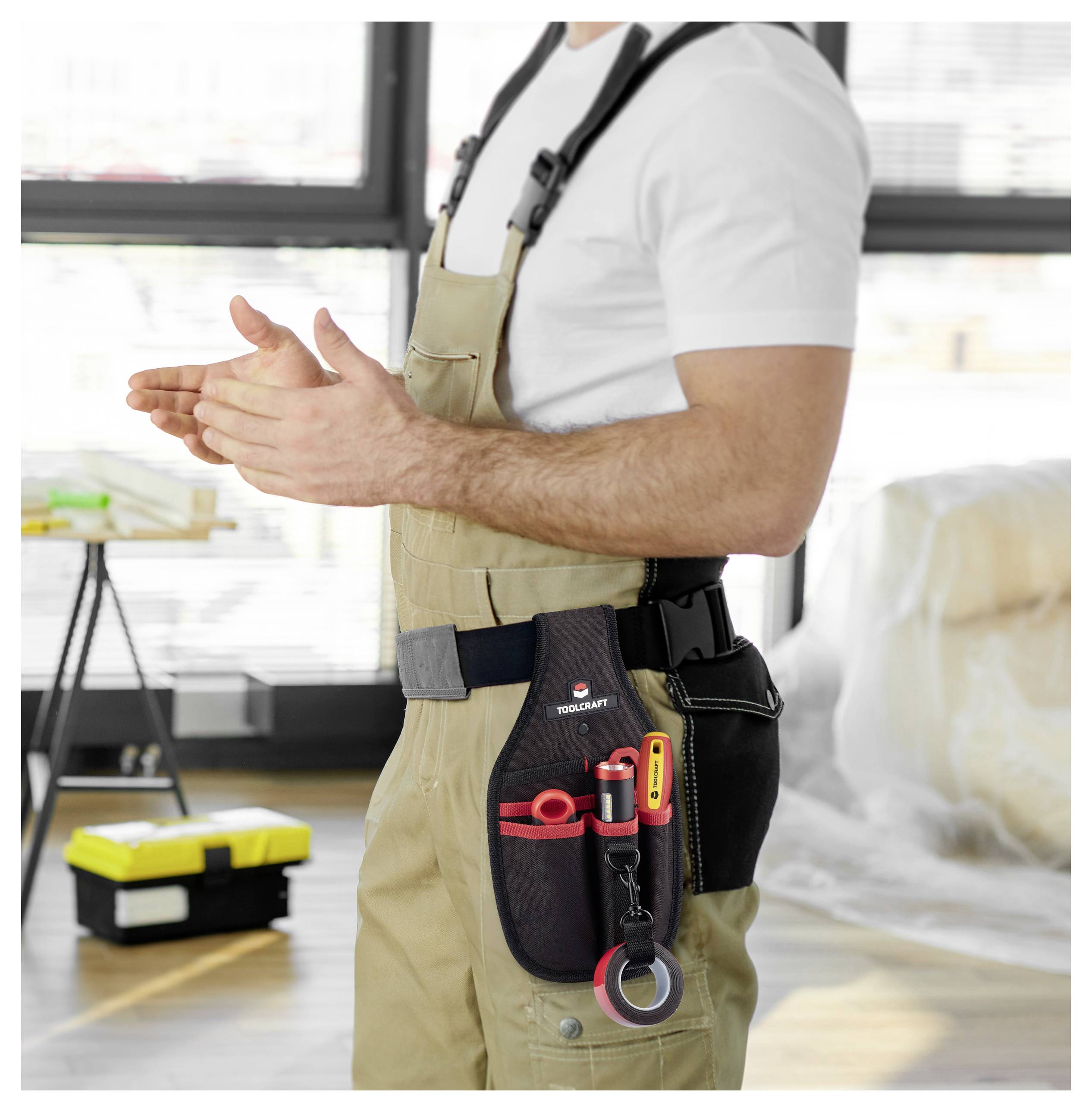 Eine Person in Overalls mit einem Werkzeuggürtel, der verschiedene Werkzeuge enthält, steht in einem Raum mit Baumaterialien und Kisten.