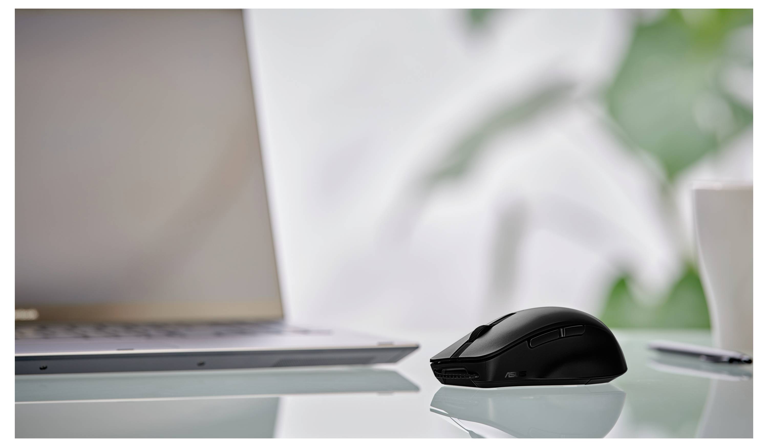 Eine schlanke schwarze kabellose Maus auf einem Glasschreibtisch neben einem teilweise sichtbaren geöffneten Laptop und einer weißen Kaffeetasse; unscharfer laubiger Hintergrund.