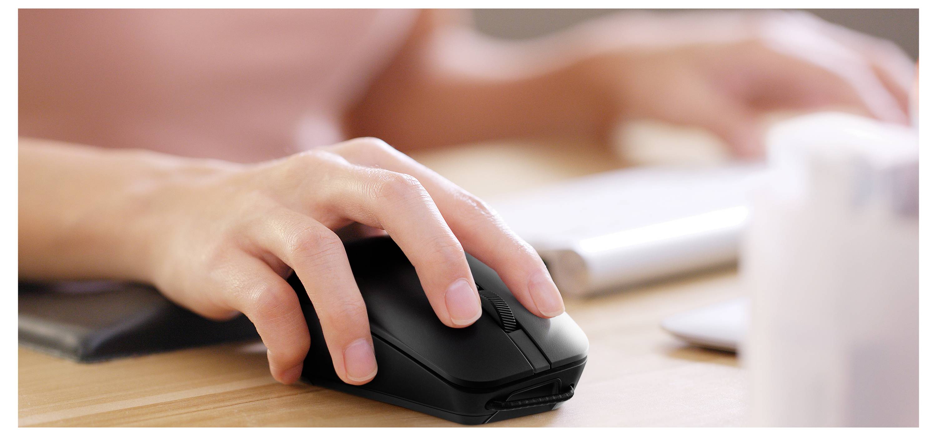 Eine Person benutzt eine schwarze Computermaus auf einem Holzschreibtisch, während im unscharfen Hintergrund Hände auf einer Tastatur tippen.
