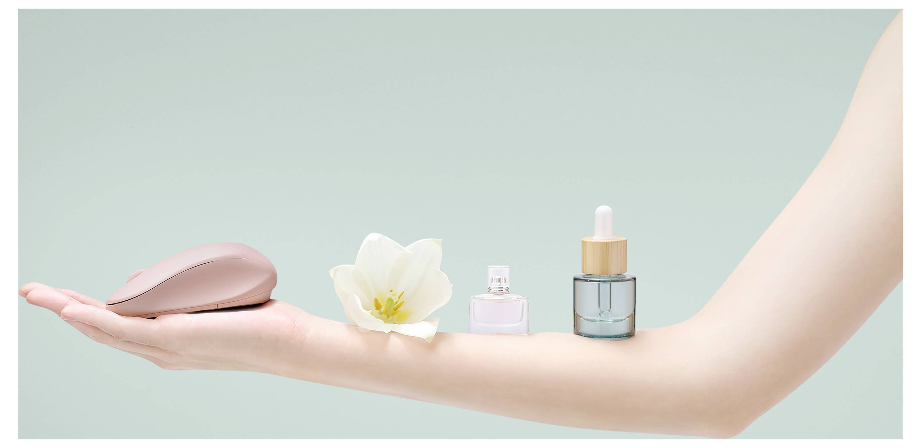 Eine ausgestreckte Hand hält eine rosa Computermaus, eine weiße Blume, kleine Parfümfläschchen und ein Serum vor einem hellgrünen Hintergrund.