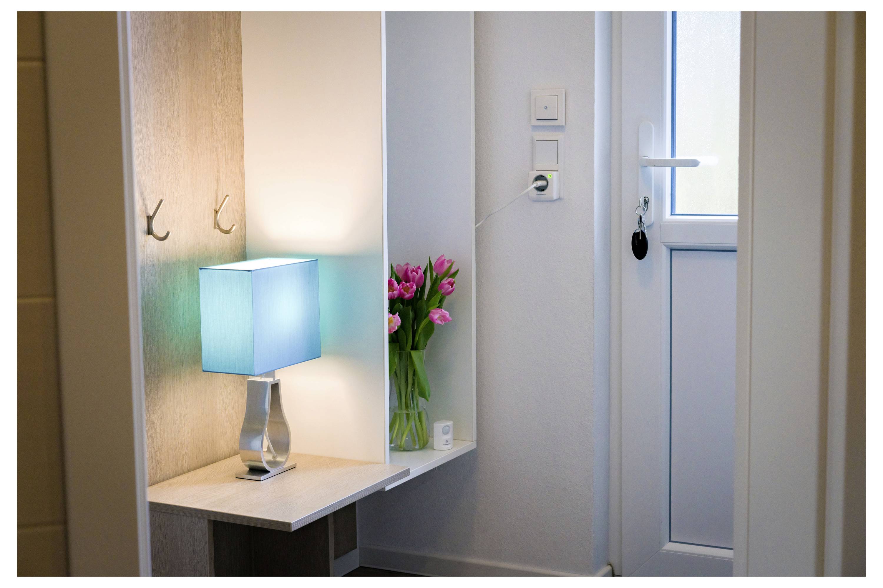 Ein kleiner Flur mit einer türkisfarbenen Lampe auf einem Ecktisch, rosa Tulpen in einer Vase und einer geschlossenen weißen Tür mit einem Schlüssel im Schloss.