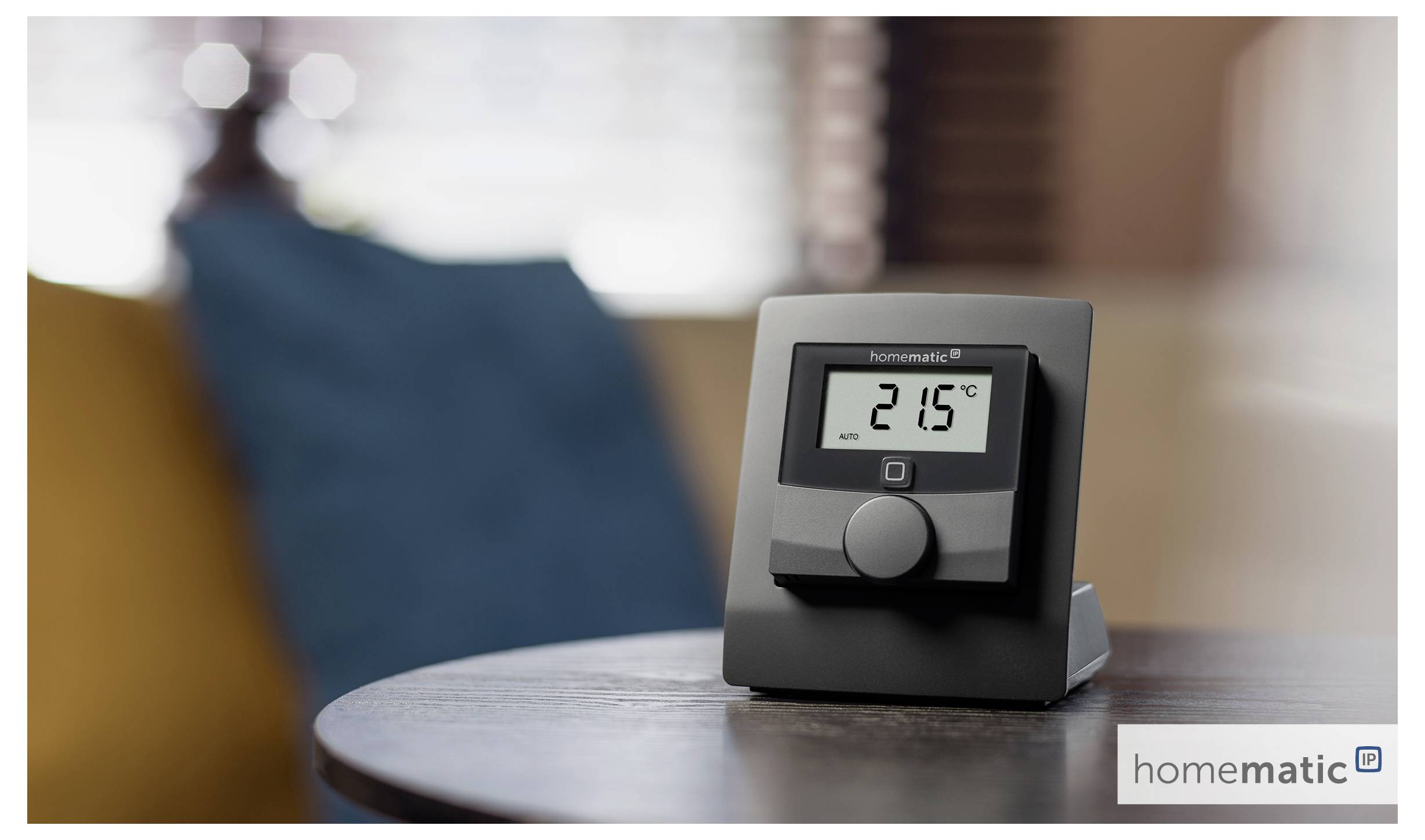 Ein digitales Thermostat auf einem Tisch zeigt '21,5°C'. Der Hintergrund umfasst verschwommene Möbel und deutet auf eine häusliche Umgebung hin.