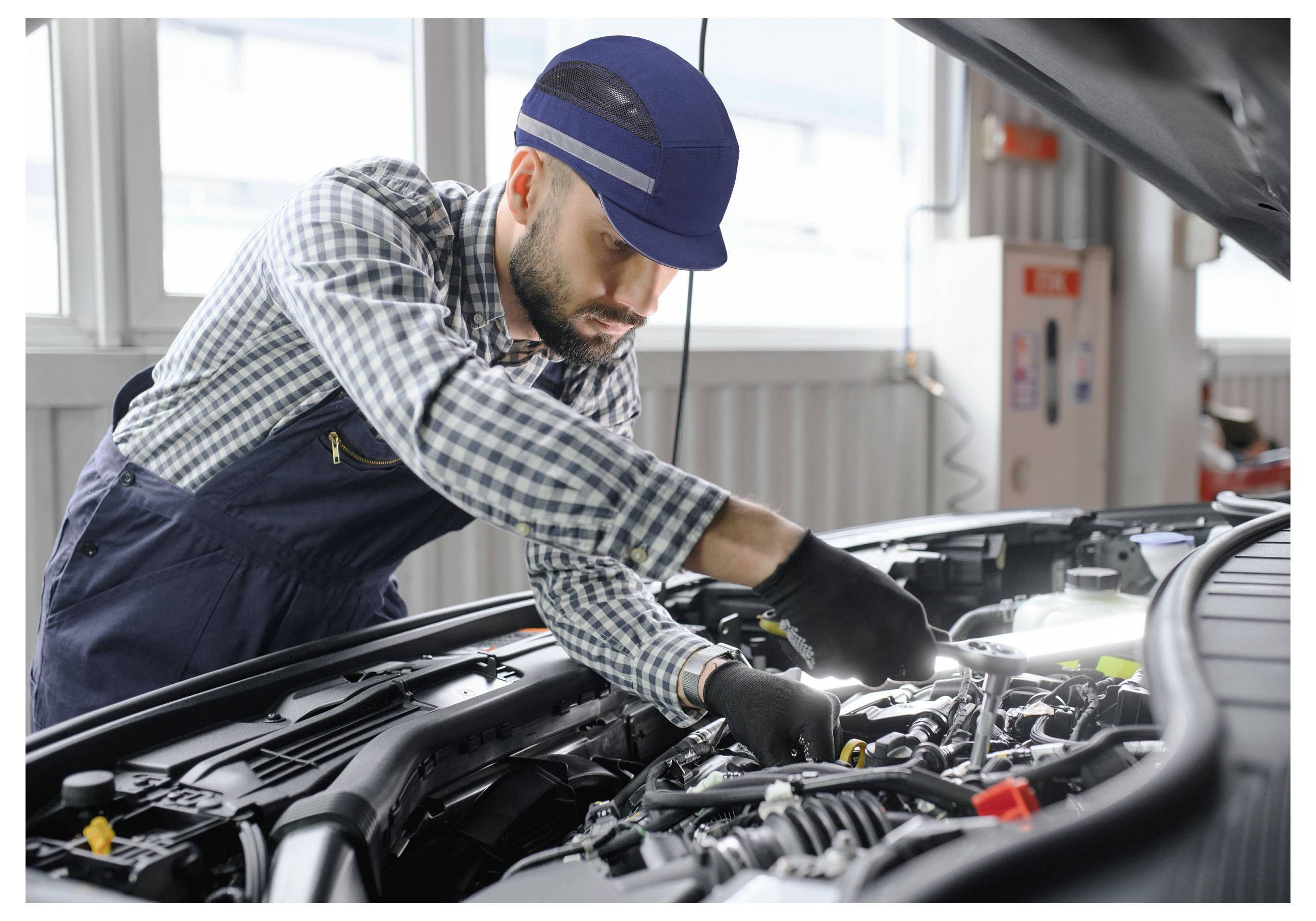 Mechaniker in einer Werkstatt repariert einen Automotor, trägt ein kariertes Hemd, eine Mütze und Handschuhe. Die Motorhaube ist offen, Werkzeuge sind sichtbar.