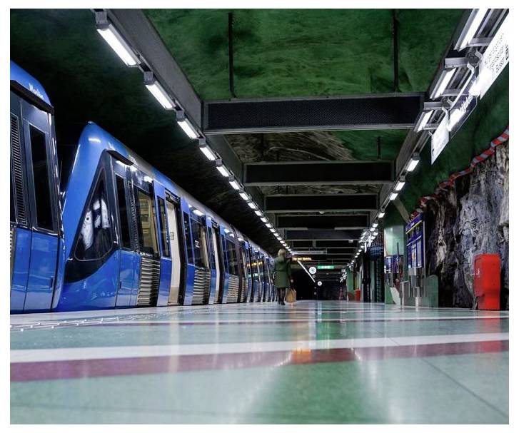 Ein blauer Zug steht an einem Bahnhof mit grünen Wänden und Boden. Eine Person in einem Mantel steht auf dem Bahnsteig. Lichter beleuchten den Tunnel.