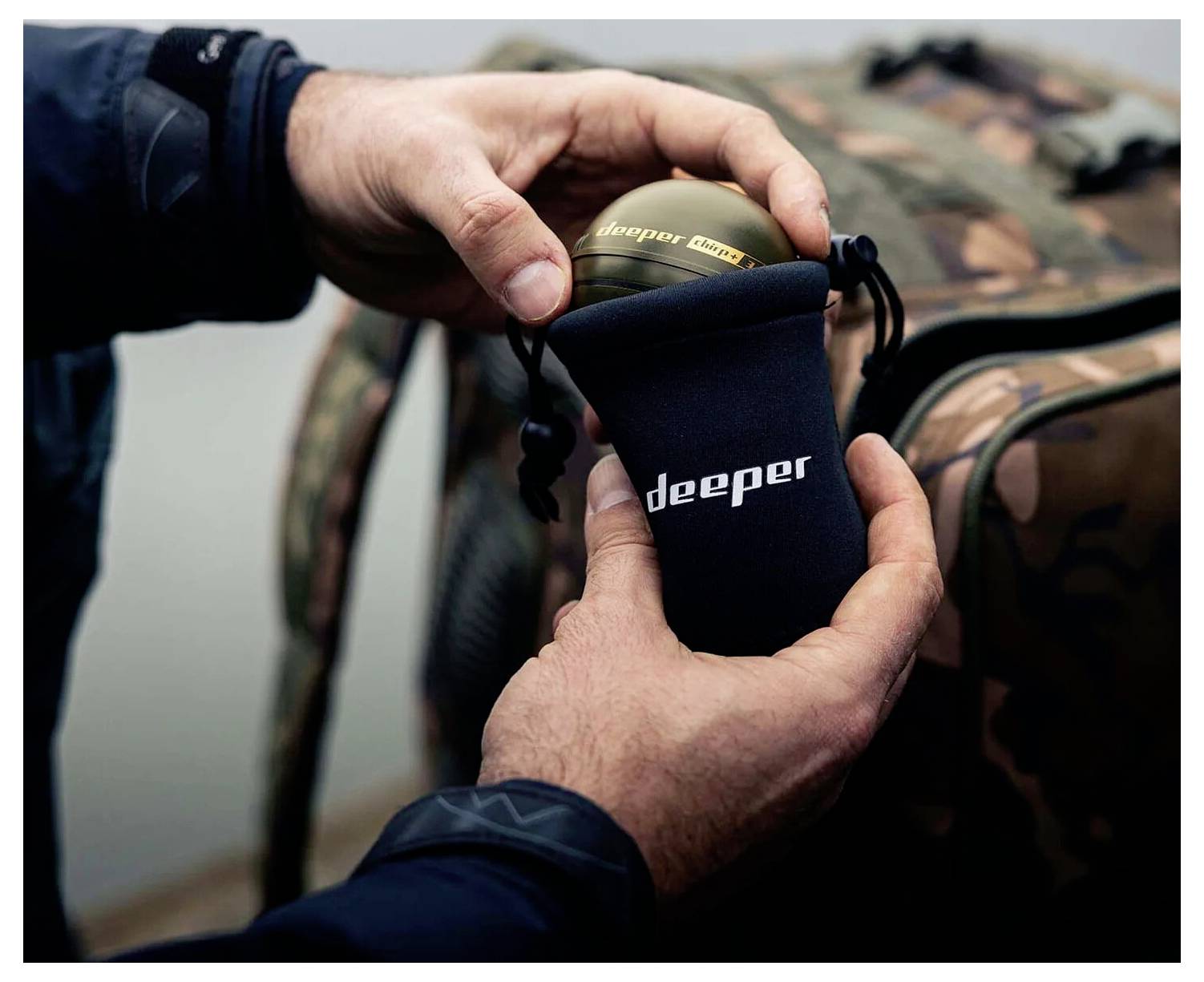 Eine Person hält ein rundes Gerät mit der Aufschrift „deeper" über einer schwarzen Tasche, wobei sich im Hintergrund eine Camouflage-Tasche befindet.