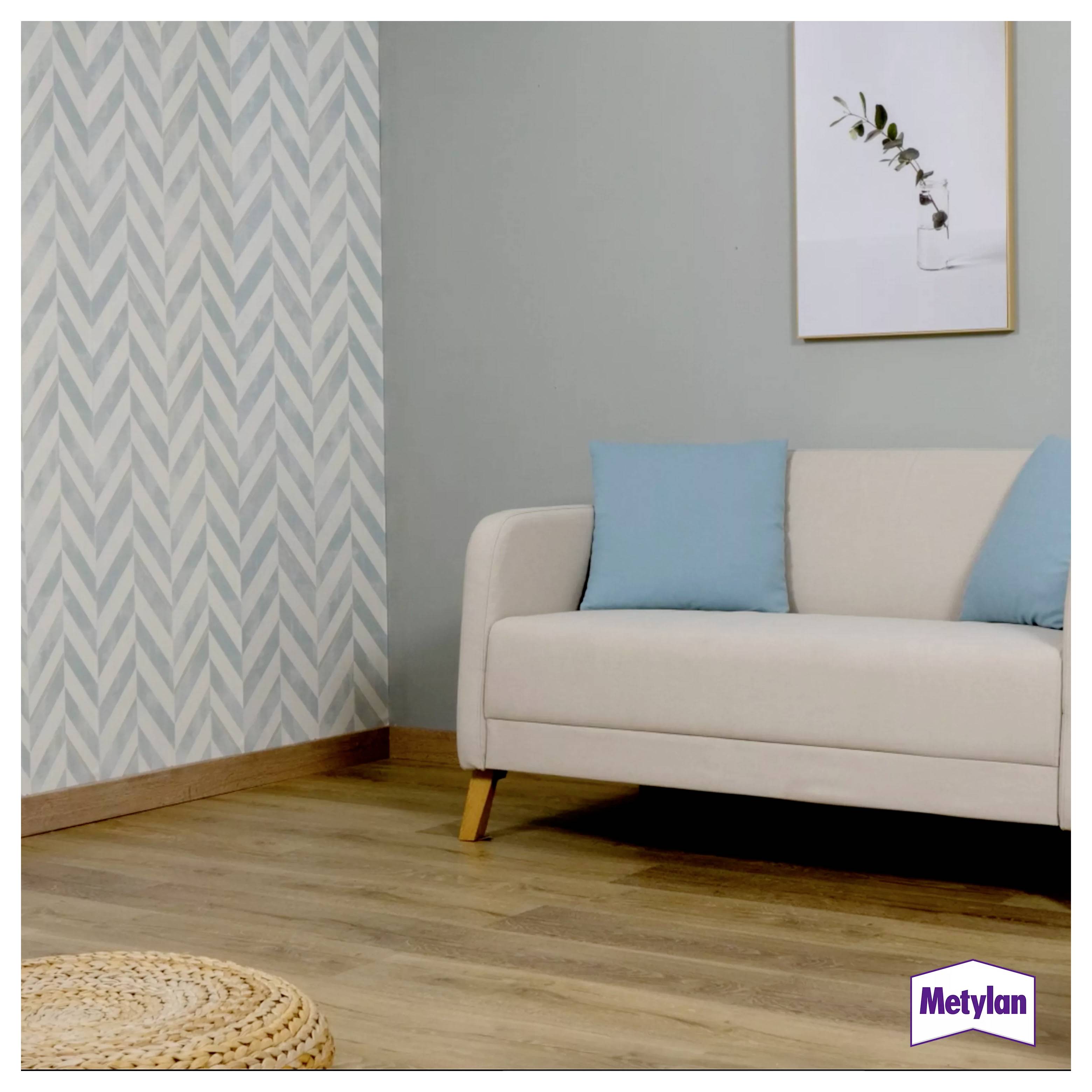 Ein moderner Wohnraum zeigt ein weißes Sofa mit blauen Kissen, gemusterte Tapeten, Holzfußboden und ein gerahmtes Kunstwerk mit Blättermotiv.