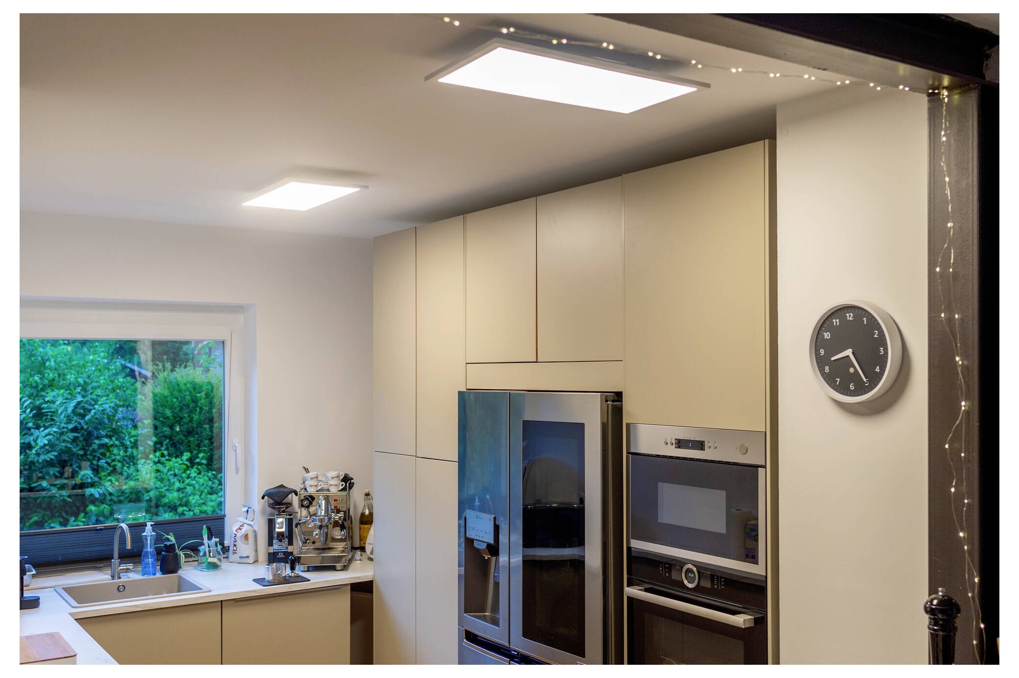 Eine moderne Küche mit beigen Schränken, einem blauen Kühlschrank und eingebauten Haushaltsgeräten. Eine Wanduhr zeigt 12:15 Uhr. Ein Fenster gibt den Blick auf Grünpflanzen im Außenbereich frei.