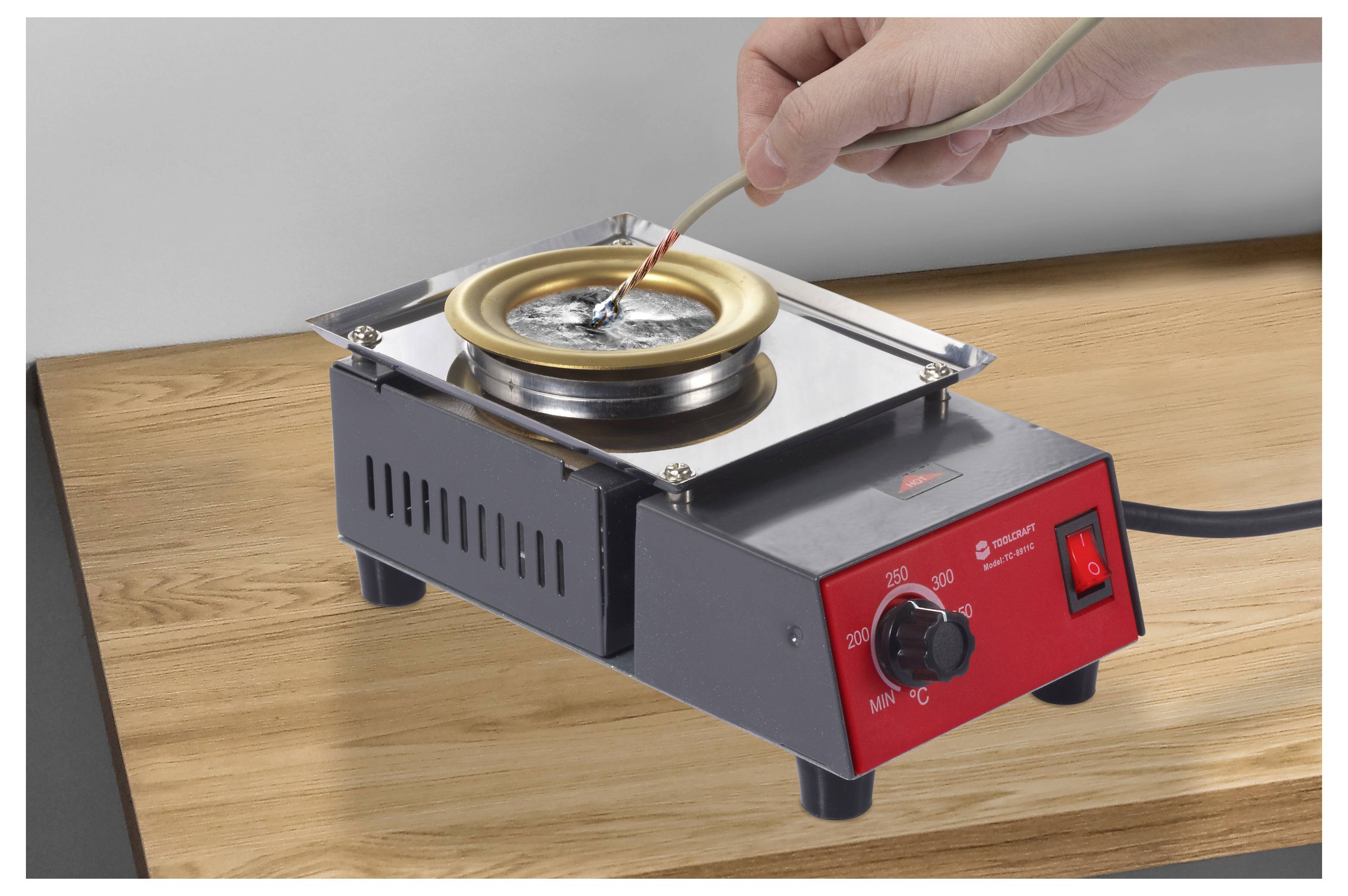 Eine Hand hält einen Draht und taucht ihn in einen kleinen elektrischen Lötopf auf einem Holztisch. Der Topf ist auf 200 Grad Celsius eingestellt.