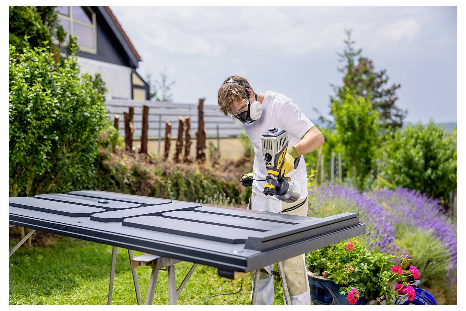 Eine Person mit Sicherheitsausrüstung verwendet ein Elektrowerkzeug, um eine große, dunkel gefärbte Holzplatte im Freien zu schneiden. Üppiges Grün umgibt das Gebiet.