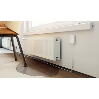 Eine kleine Wasserpfütze auf einem Holzboden deutet auf einen Heizkörperleck hin; ein weißer Sensor ist in der Nähe zur Überwachung angebracht.