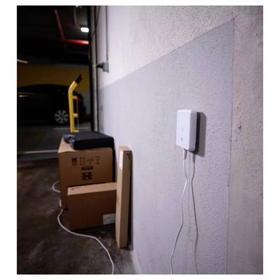 Kisten und ein schwarzer Koffer sind an einer Wand eines Parkhauses gestapelt, wobei ein Gerät an der Wand montiert ist und durch ein weißes Kabel verbunden ist.