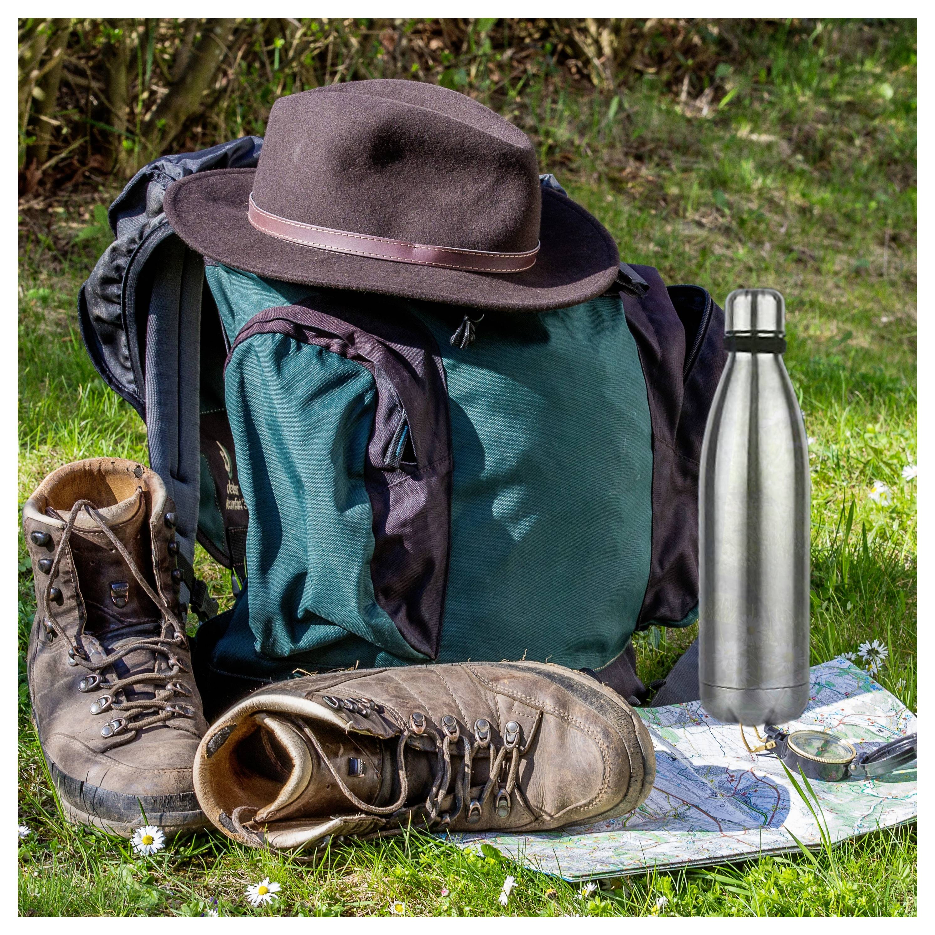 Ein grüner Rucksack mit einem braunen Hut obenauf, Wanderschuhe, eine Karte und eine Metallflasche auf Gras, die ein Outdoor-Abenteuer andeuten.