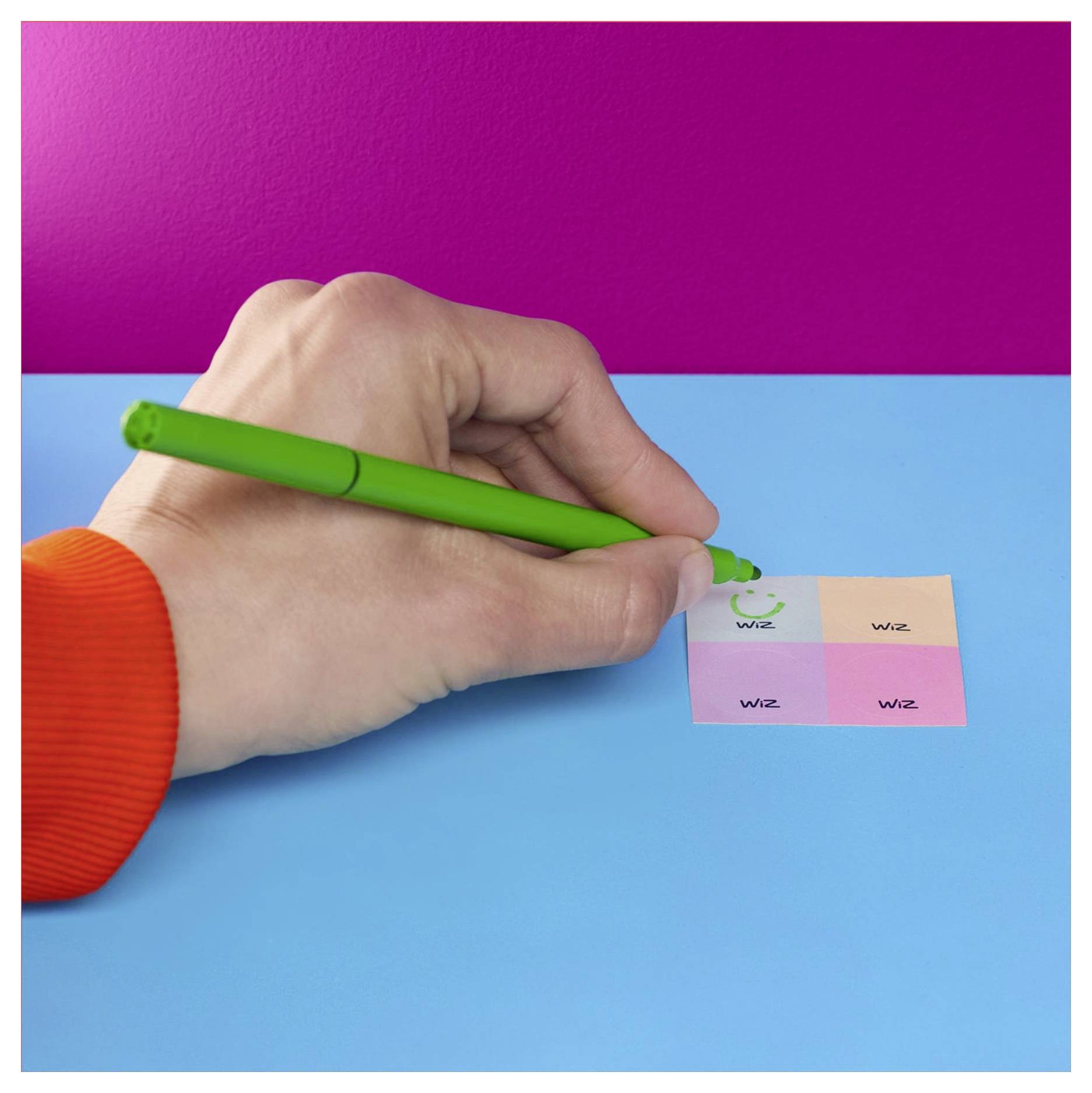 Eine Hand in einem orangefarbenen Ärmel hält einen grünen Stift über einem Farbprüfstreifen auf einer blauen Oberfläche mit einem pinken Hintergrund.