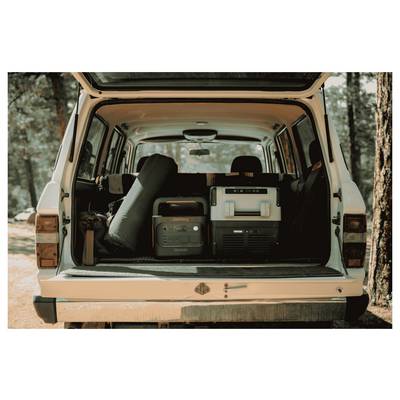 Die Rückseite eines Fahrzeugs ist geöffnet und zeigt Campingausrüstung, darunter einen Kühlschrank, eine Powerstation und einen zusammengerollten Schlafsack, vor einer Waldkulisse.