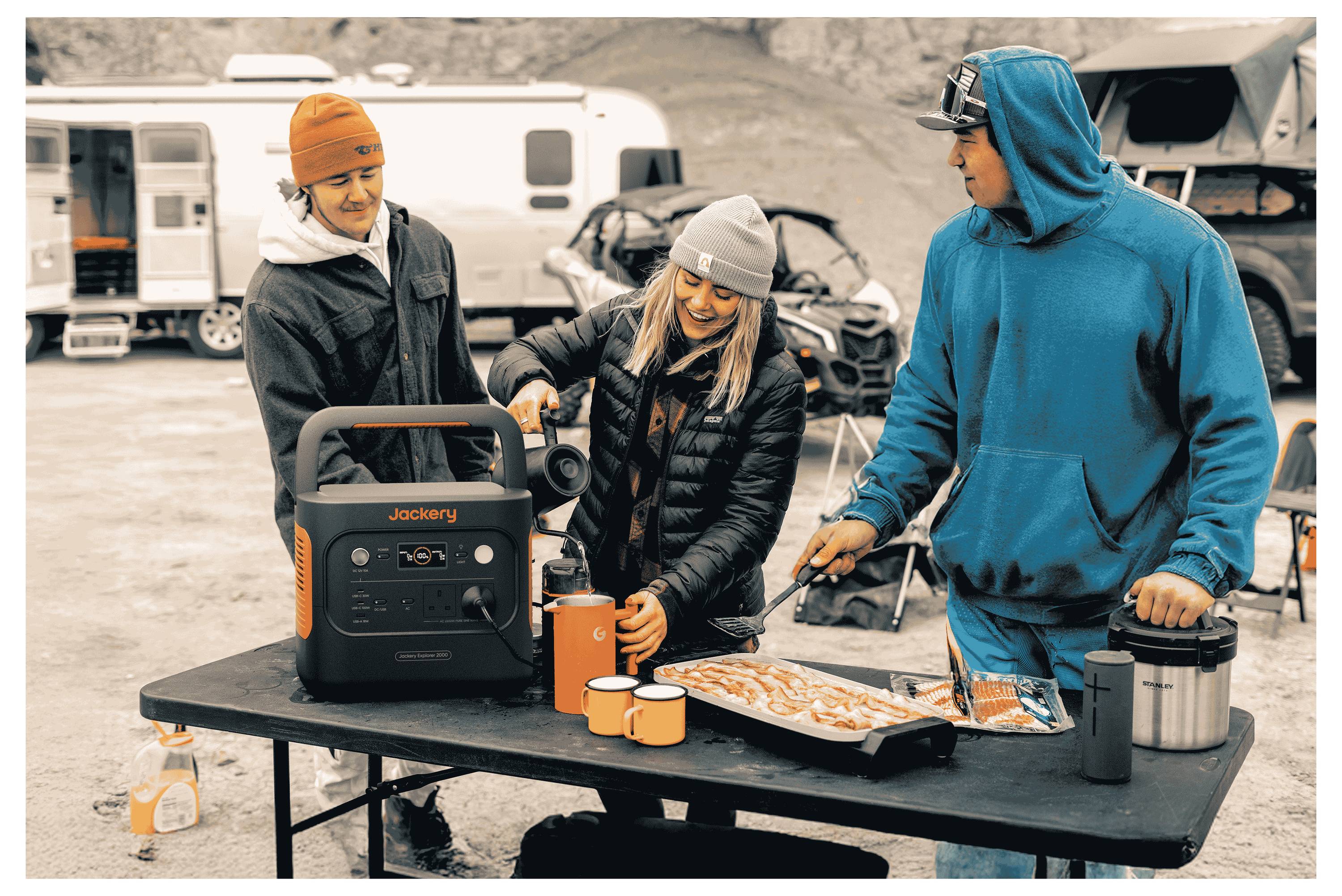 Menschen im Freien, die Essen an einem Tisch mit einer mobilen Stromstation, einem Grill und Bechern zubereiten. Fahrzeuge und Zelte sind im Hintergrund zu sehen.