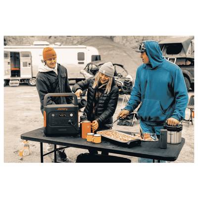Menschen im Freien, die Essen an einem Tisch mit einer mobilen Stromstation, einem Grill und Bechern zubereiten. Fahrzeuge und Zelte sind im Hintergrund zu sehen.