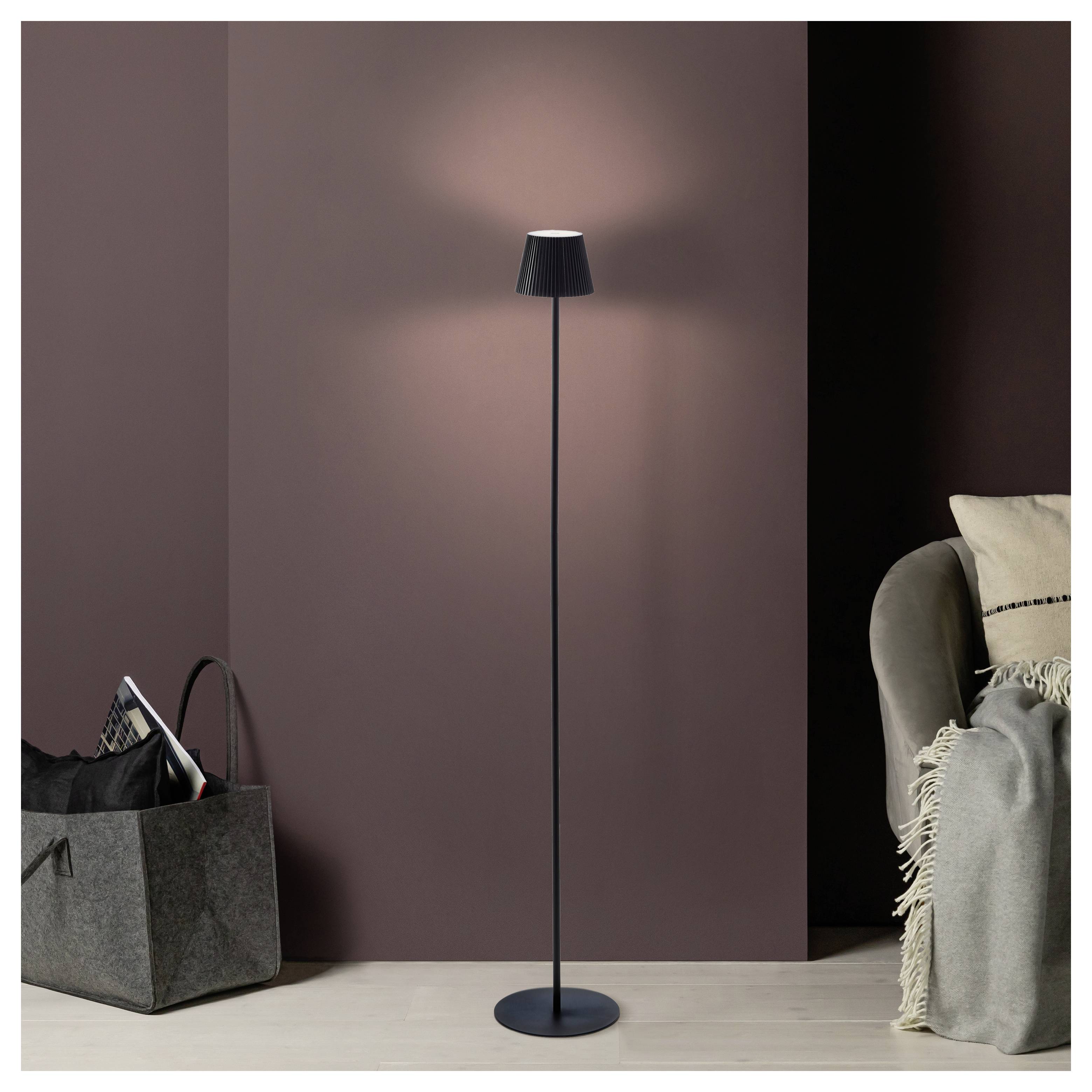 Eine hohe, schlanke Stehlampe mit einem schwarzen Lampenschirm beleuchtet eine gemütliche Ecke und hebt einen grauen Sessel mit einer gefransten Decke und einem Korb hervor.