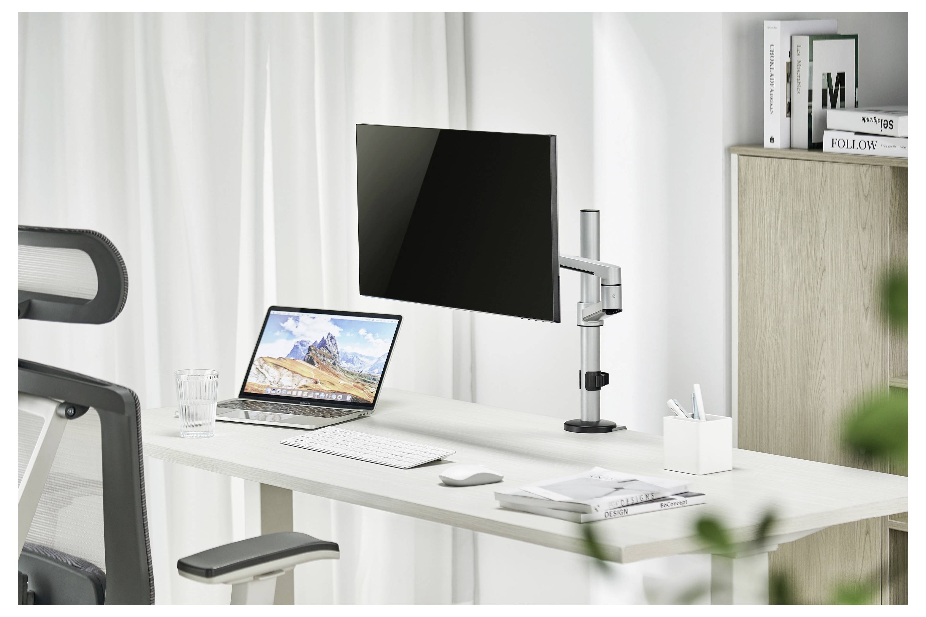 Ein modernes Büro-Setup mit einem aufgeklappten Laptop, der eine Berglandschaft zeigt, einem an der Wand montierten Monitor, einer Tastatur, einer Maus und minimalistischer Dekoration.