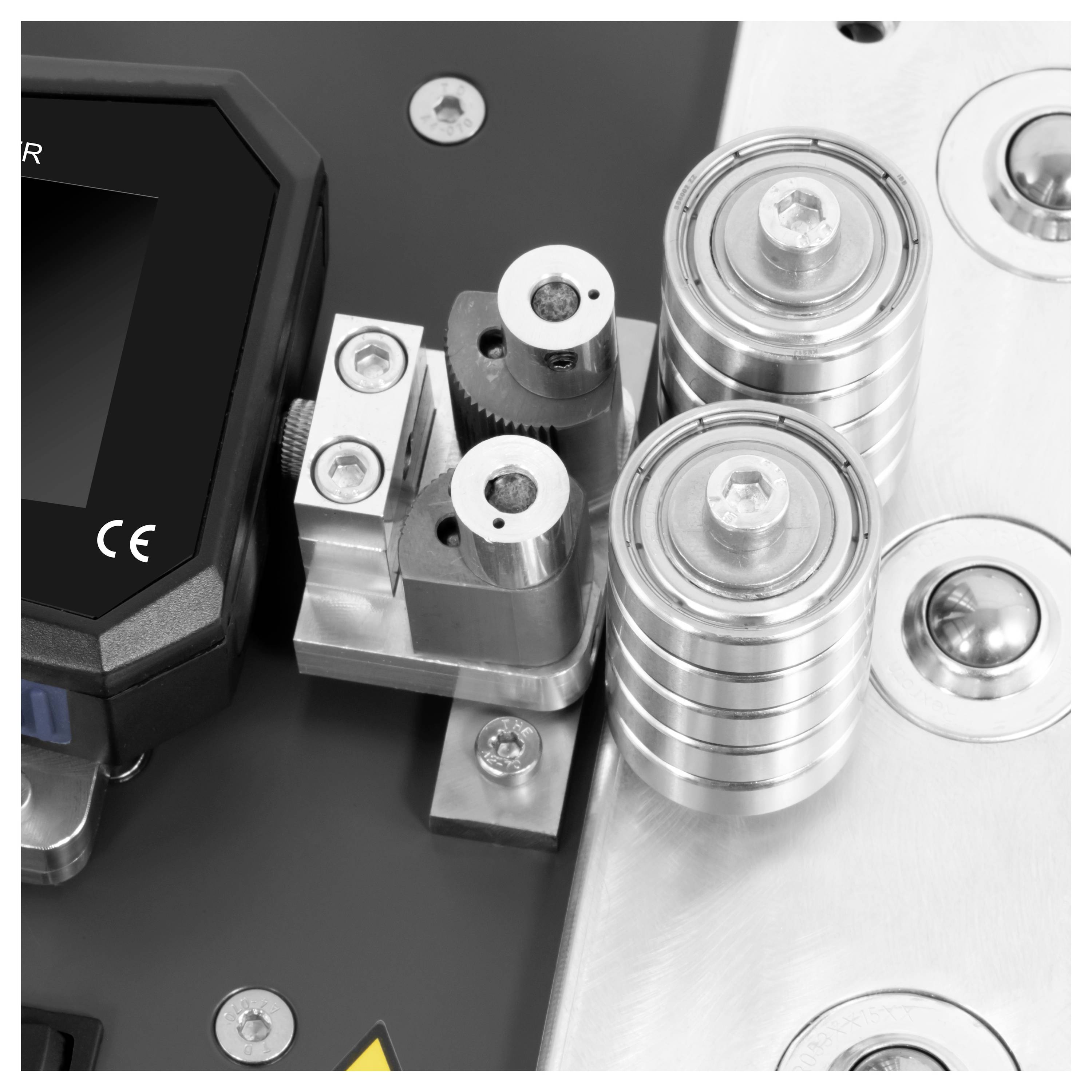 Nahaufnahme einer mechanischen Baugruppe mit Metallrollen, Halterungen und einem schwarzen Gerätekörper, der auf einer silbernen Grundplatte montiert ist.
