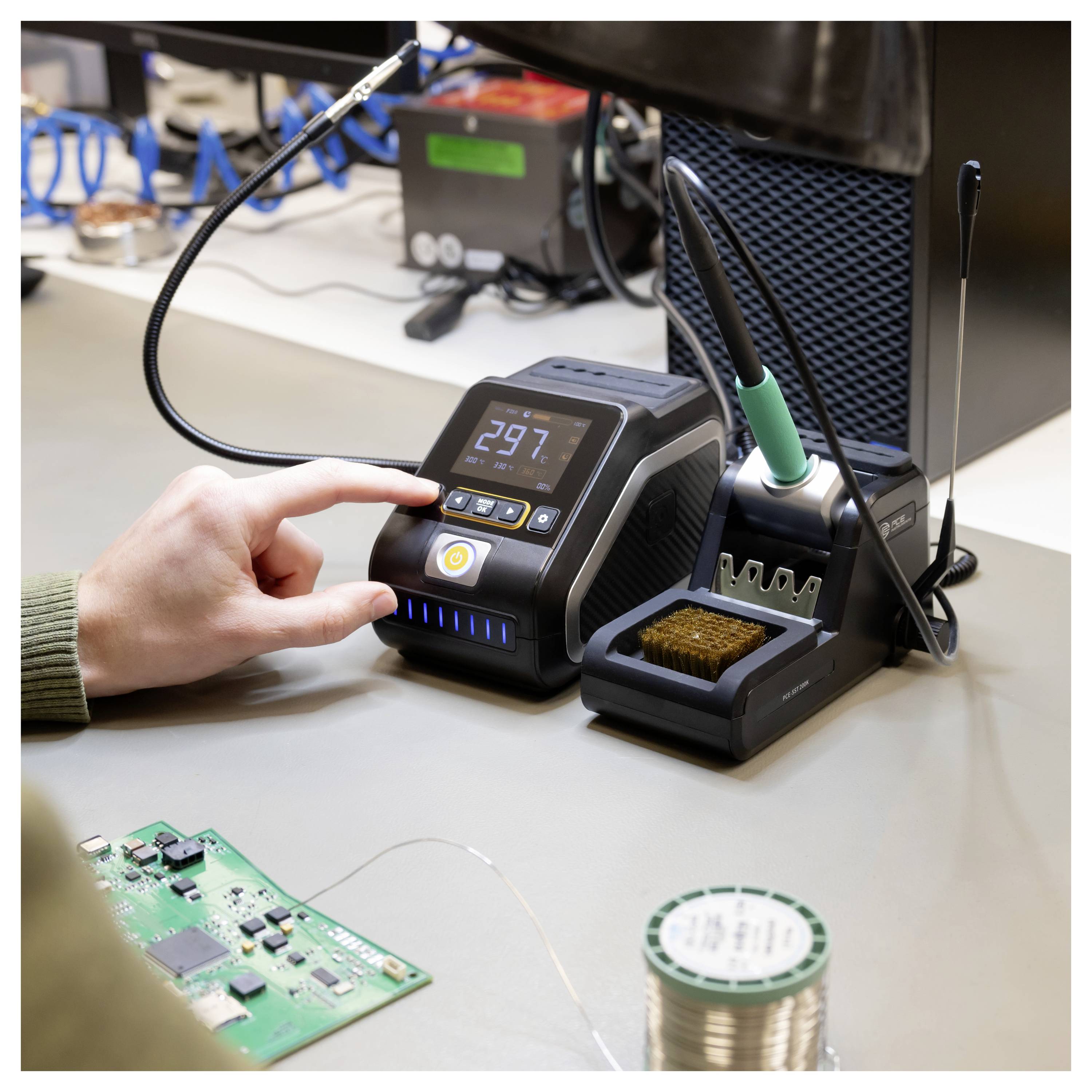 Hand stellt eine digitale Lötstation ein, die 297°C anzeigt, mit einem Lötkolben in seinem Ständer neben einer Elektronikwerkbank und einer Leiterplatte.