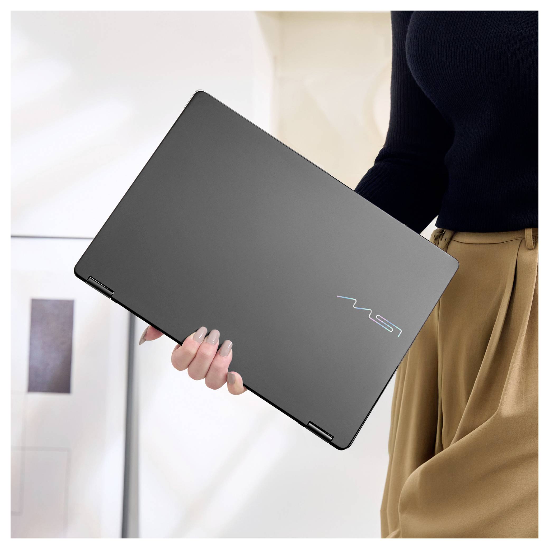 Eine Person in einem schwarzen Pullover hält einen geschlossenen schwarzen Laptop mit einem farbenfrohen Logo und steht in einem Innenraum mit unscharfem Hintergrund.