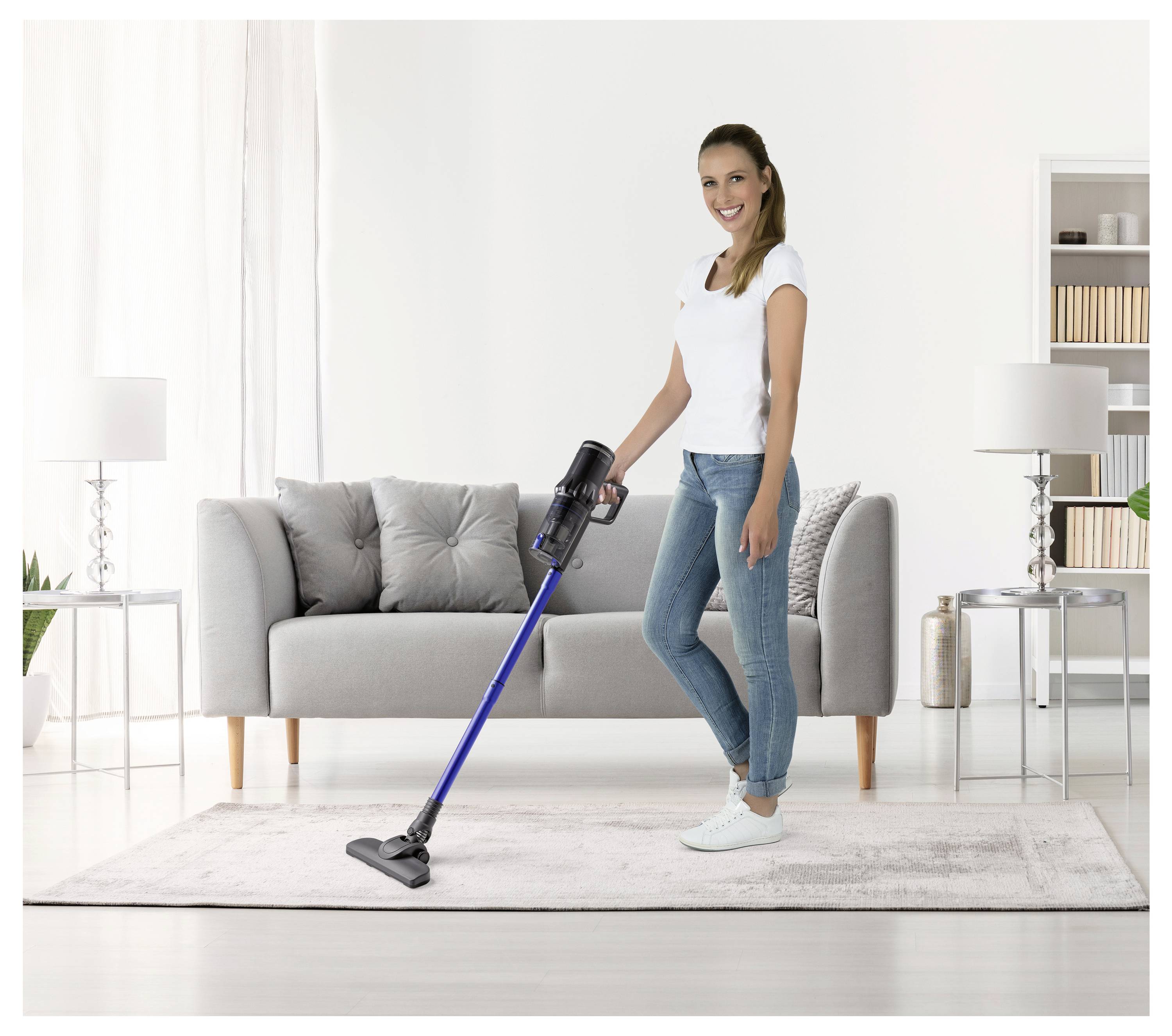 Eine Person saugt einen hellfarbigen Teppich in einem modernen Wohnzimmer mit einem grauen Sofa, zwei Lampen und einem Regal mit Büchern.