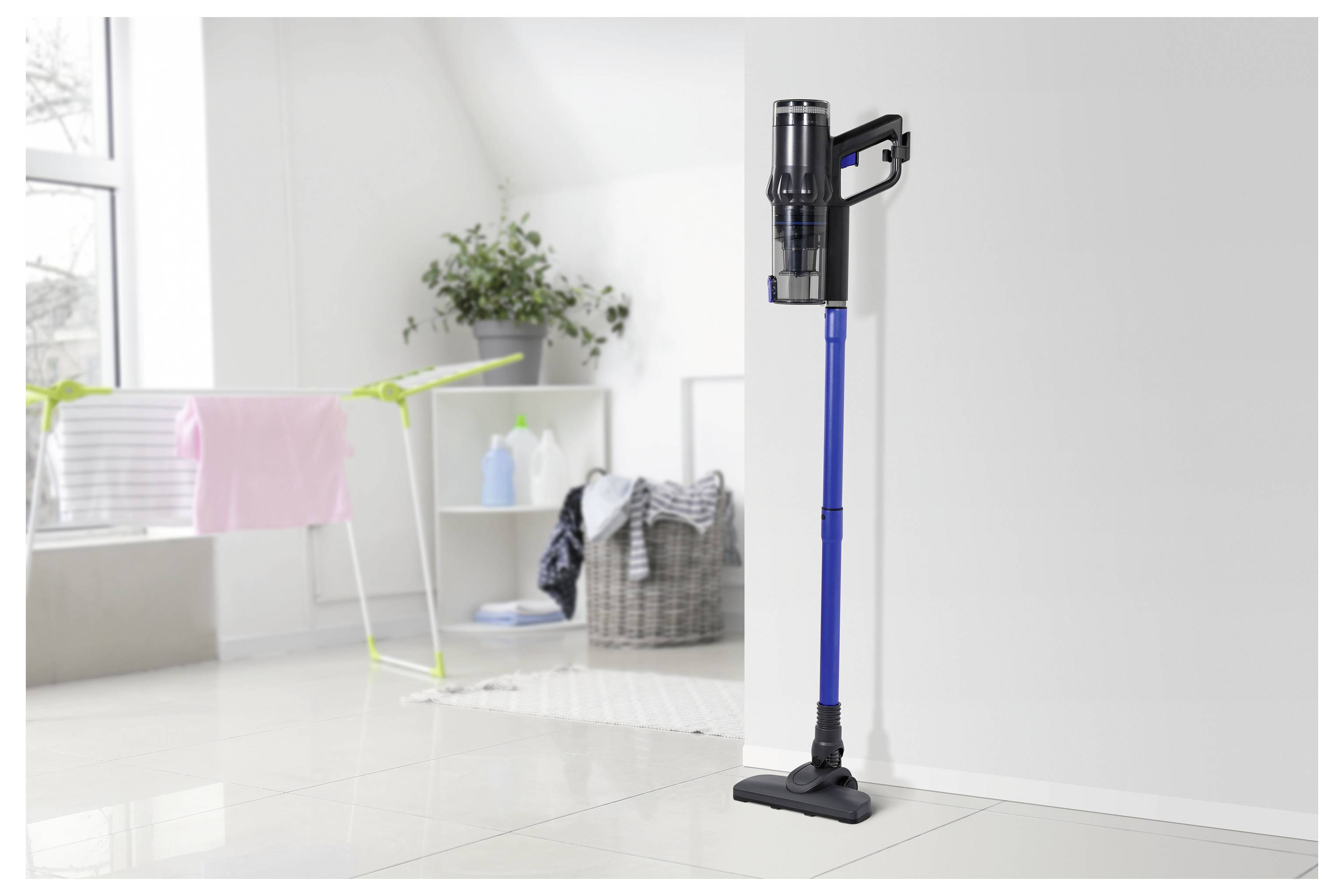 Ein kabelloses Stabstaubsaugergerät mit blauem Griff steht in einem hellen Waschraum neben einem Wäscheständer und einem Wäschekorb.