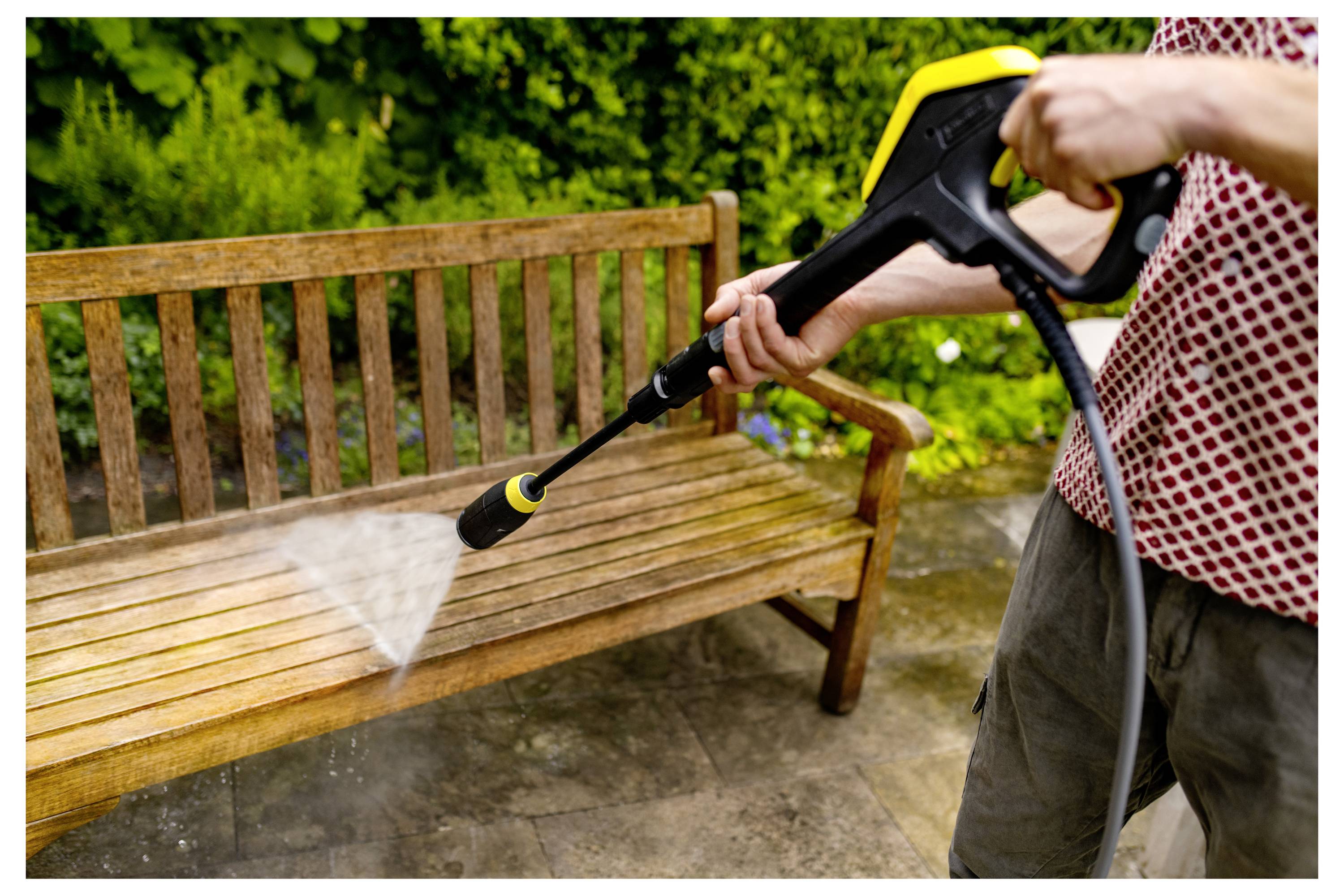 Eine Person nutzt einen Hochdruckreiniger, um eine hölzerne Gartenbank im Freien zu reinigen, wobei Wasser auf die Latten der Bank spritzt.