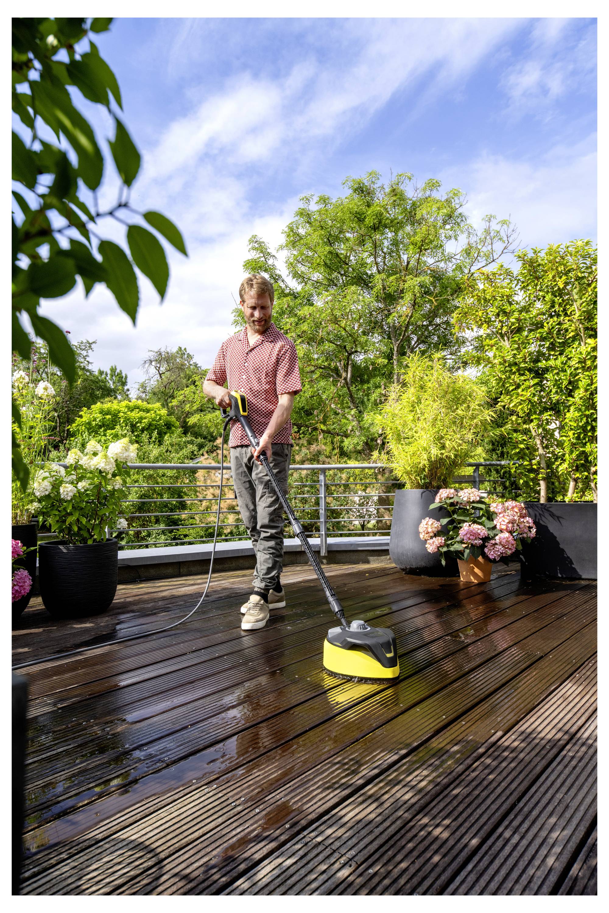 Ein Mann reinigt ein Holzdecks mit einem Hochdruckreiniger an einem sonnigen Tag, umgeben von Pflanzen und grünem Bewuchs.