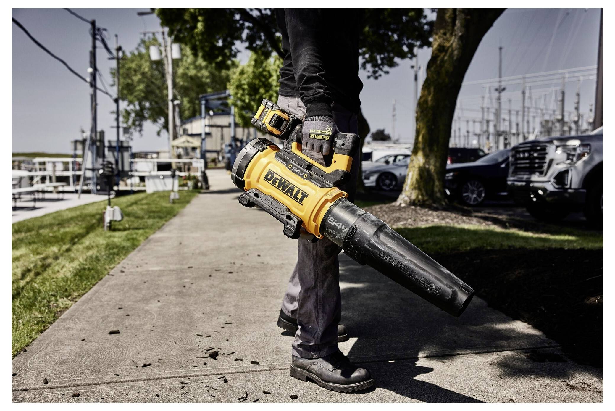 Eine Person geht auf einem Gehweg und trägt ein gelb-schwarzes Dewalt-Laubgebläse, wobei geparkte Autos und Bäume im Hintergrund sichtbar sind.